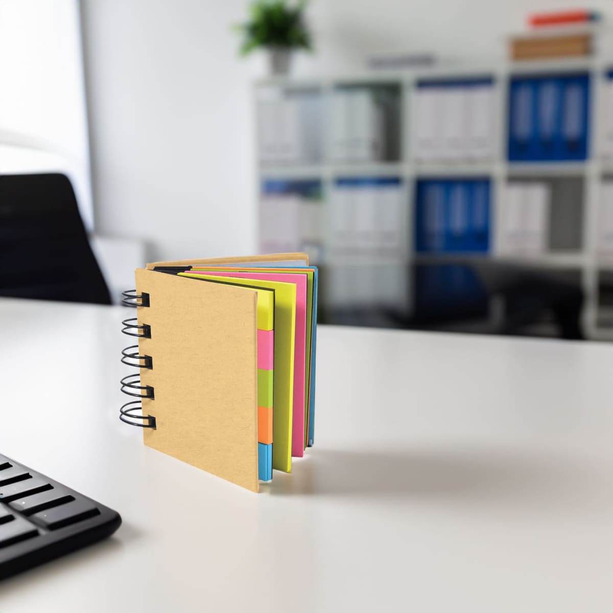 A notepad with a brown cover and colourful pages, placed on a desk in an office setting. It has a spiral binding.