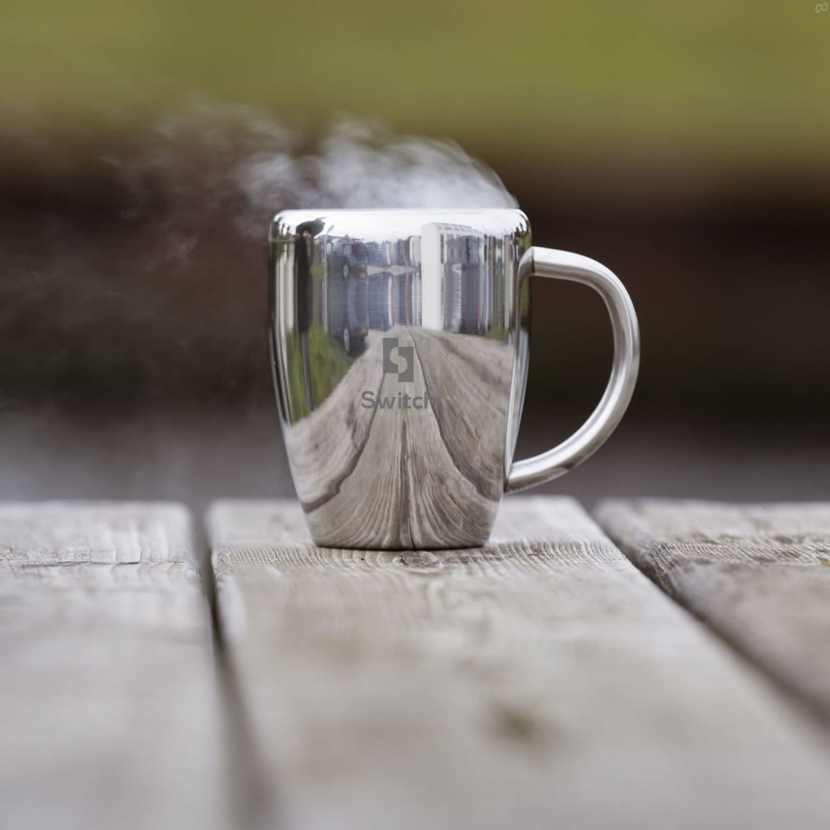 A silver coffee mug with a reflective surface and steam rising, placed on a wooden table. It features a logo.