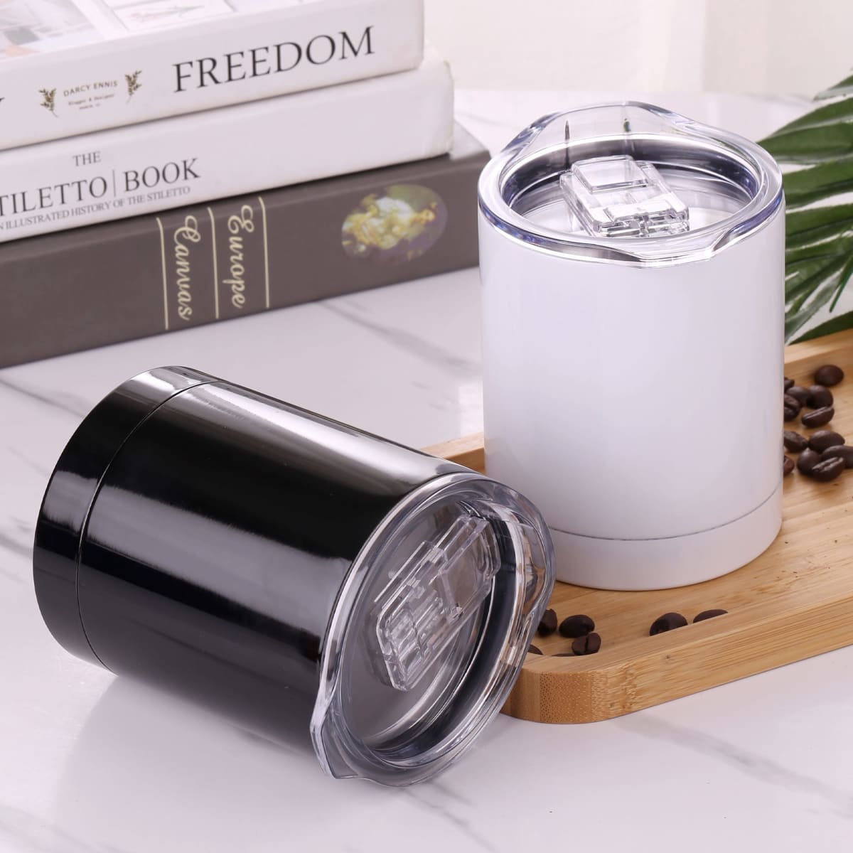 A pair of coffee mugs in black and white, featuring transparent lids, placed on a wooden tray with coffee beans.