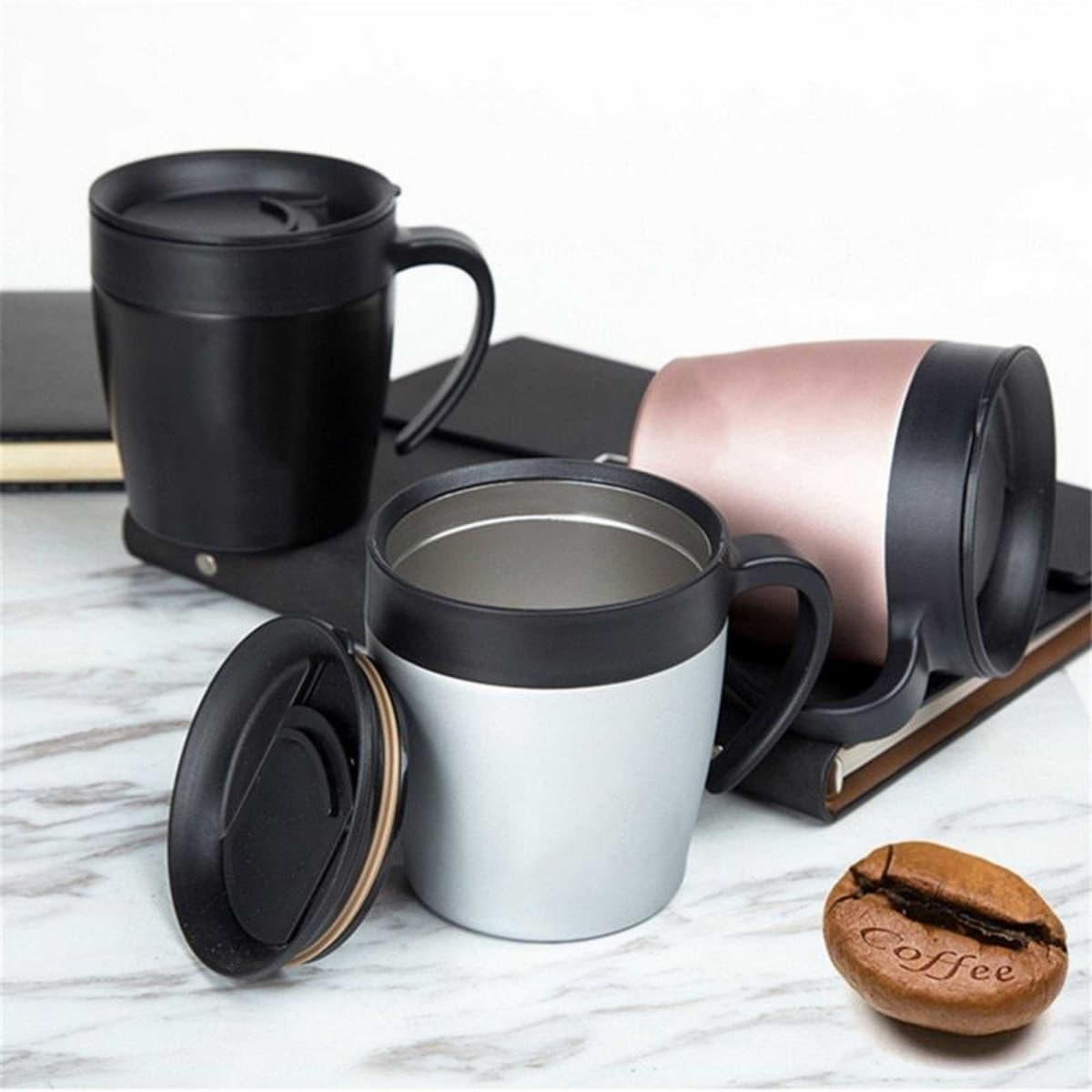 A set of three Boston coffee mugs in black, silver, and rose gold, with lids and handles, beside a coffee bean.