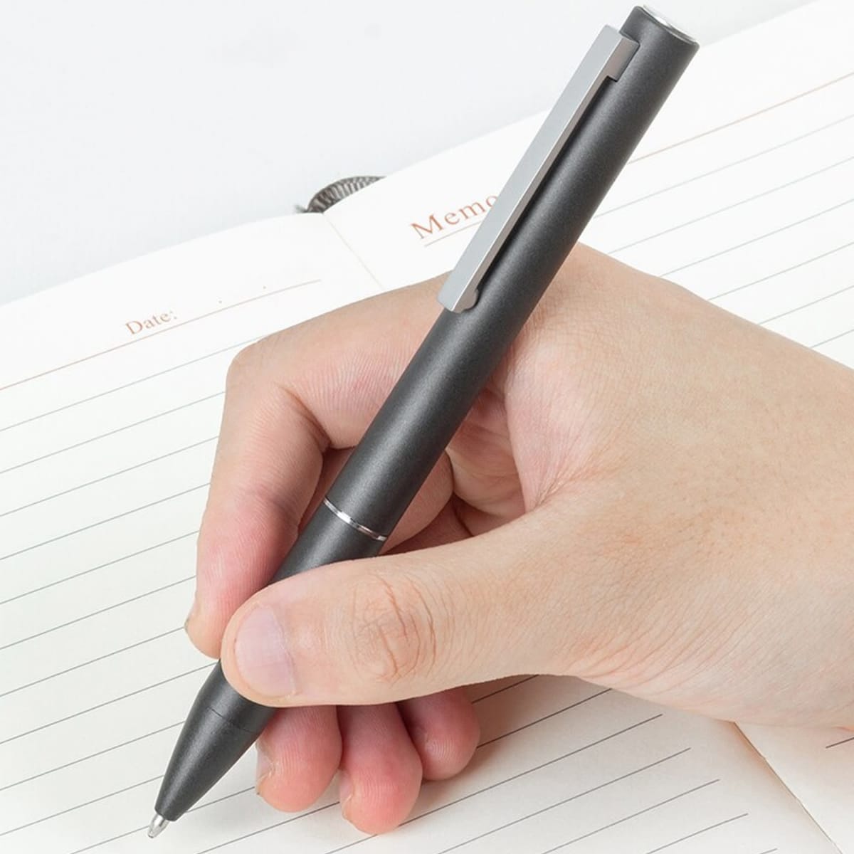 A metal pen in dark grey, held in a hand over an open notebook with lined pages.
