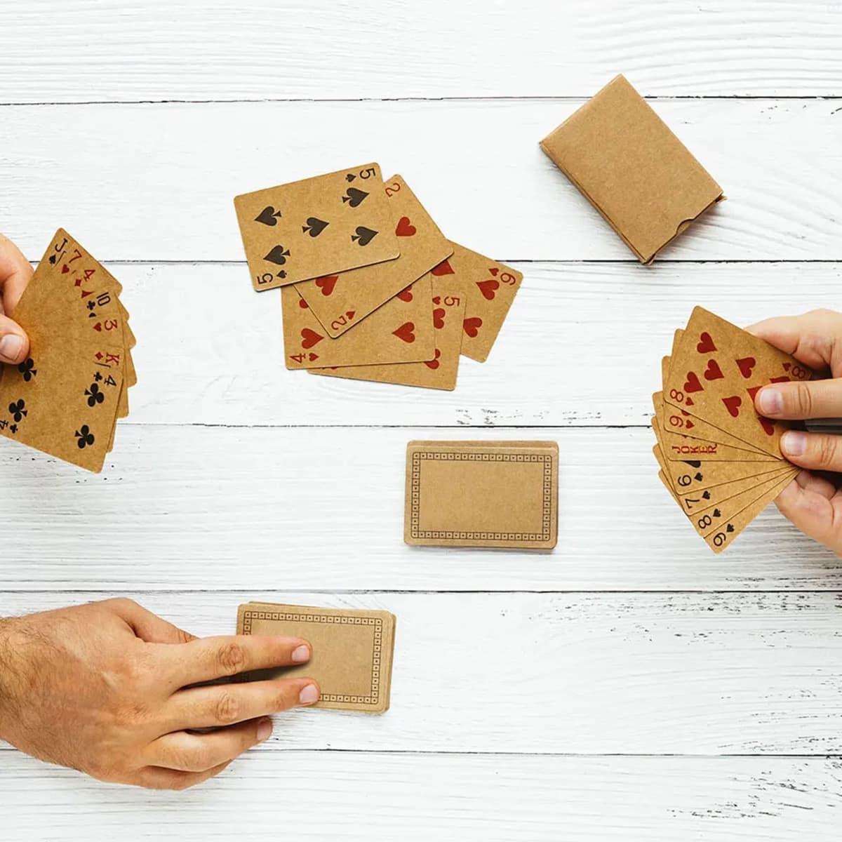 Eco-friendly playing cards in brown recycled material, displayed in hands with a deck and some cards laid out on a wooden surface.