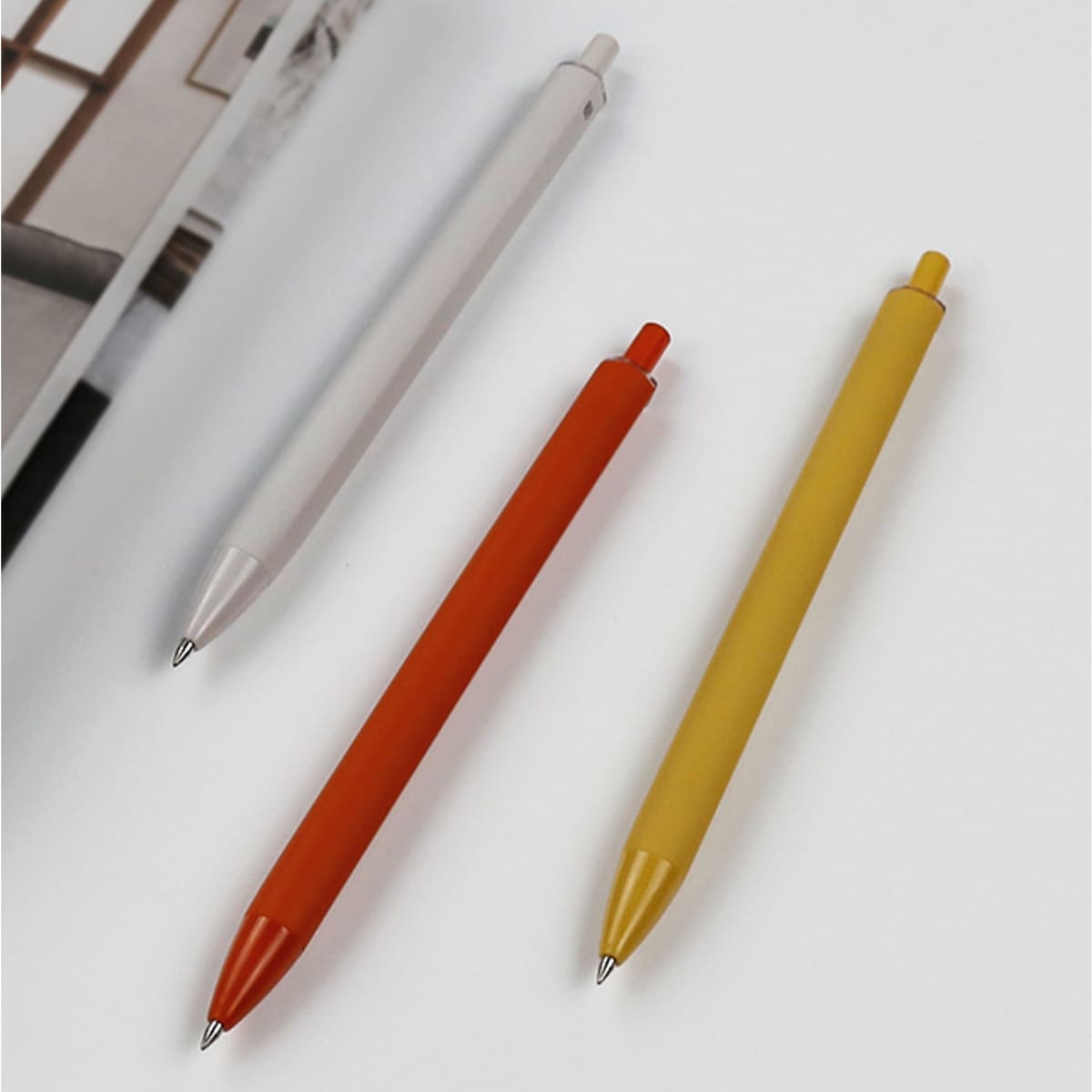 Three plastic pens in white, orange, and yellow, arranged on a flat surface next to a book. Each pen features a logo.