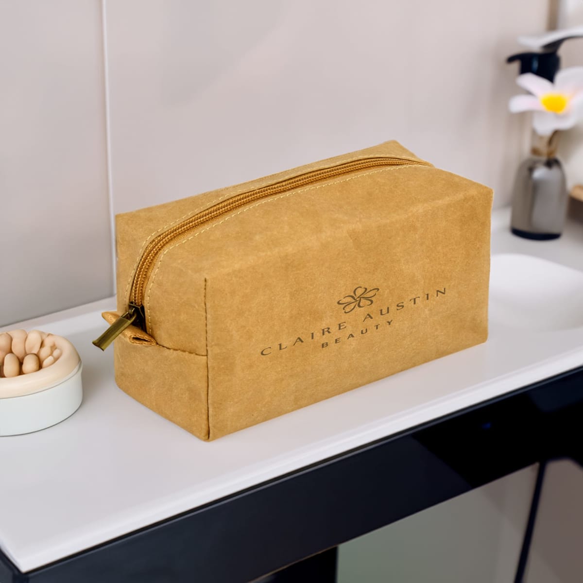 A tan kraft paper cosmetic bag with a zipper, featuring a logo, displayed on a bathroom countertop.