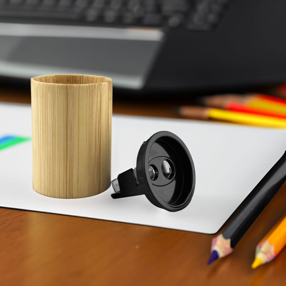A bamboo pencil sharpener with a black mechanism, placed on a desk near coloured pencils and a notepad.