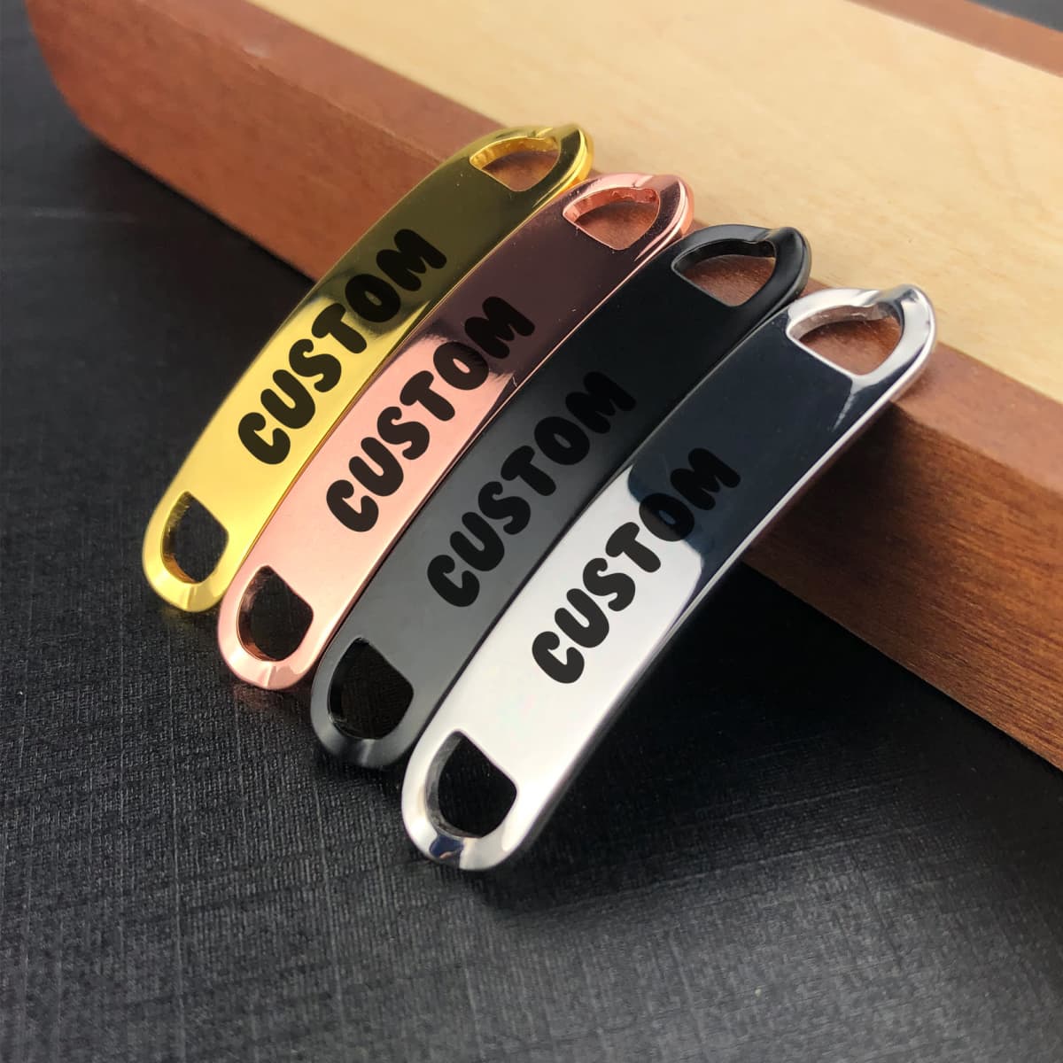 Four Smart Shoelace Charms in gold, rose gold, black, and silver, featuring a logo, arranged on a wooden surface.