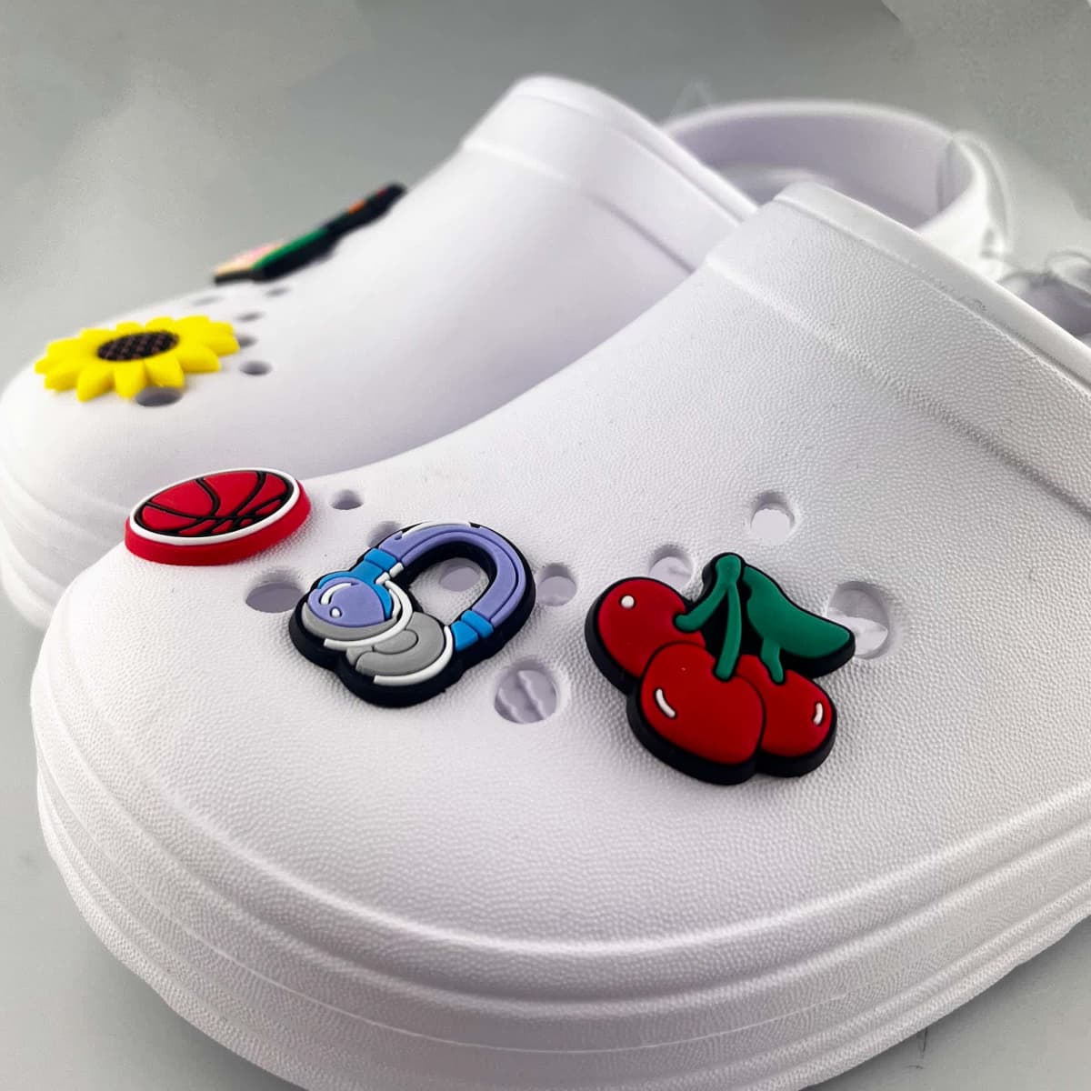 A pair of white clogs adorned with colourful Jibbits shoe charms featuring a sunflower, basketball, cherries, and a blue letter.