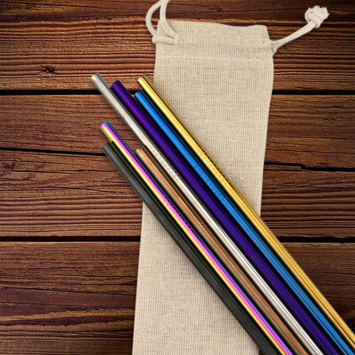 A collection of stainless steel drinking straws in various colours, resting on a canvas bag against a wooden background.