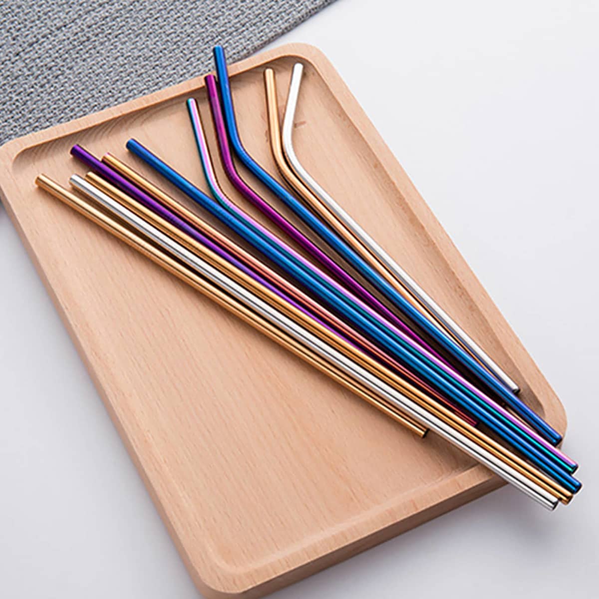 A wooden tray displaying various stainless steel drinking straws in vibrant colours, including gold, blue, purple, and silver.
