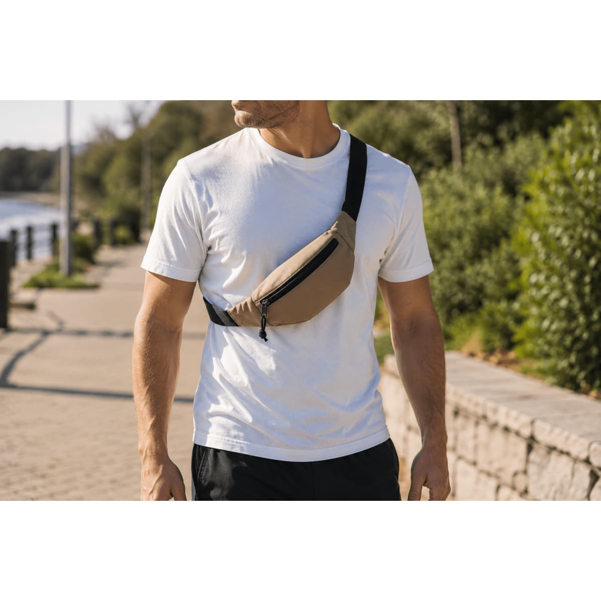 A sling bag in a tan hue worn across the chest of a person in a casual setting, with greenery and a path in the background.
