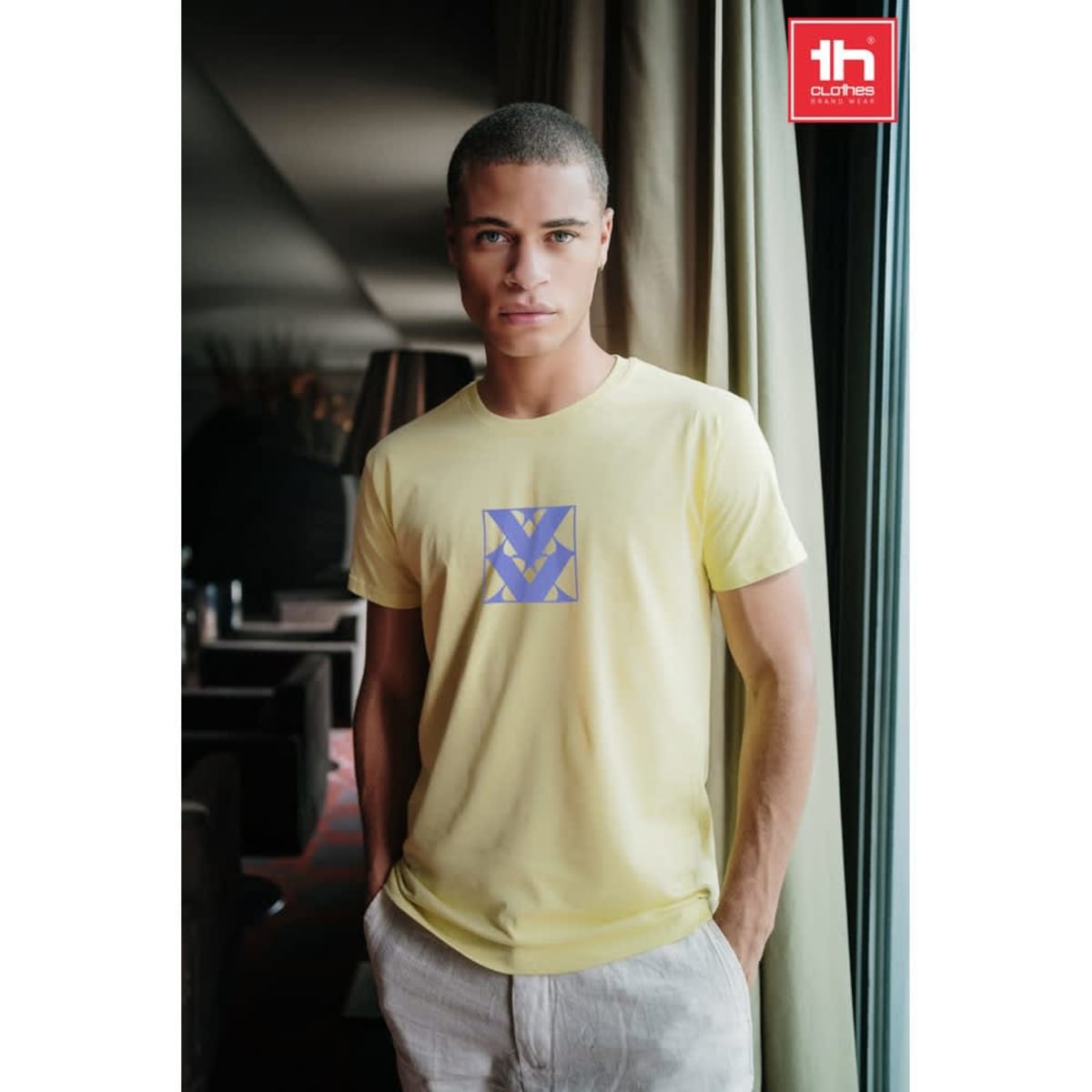 A men's tubular cotton T-shirt in yellow with a logo on the front, worn by a model indoors.