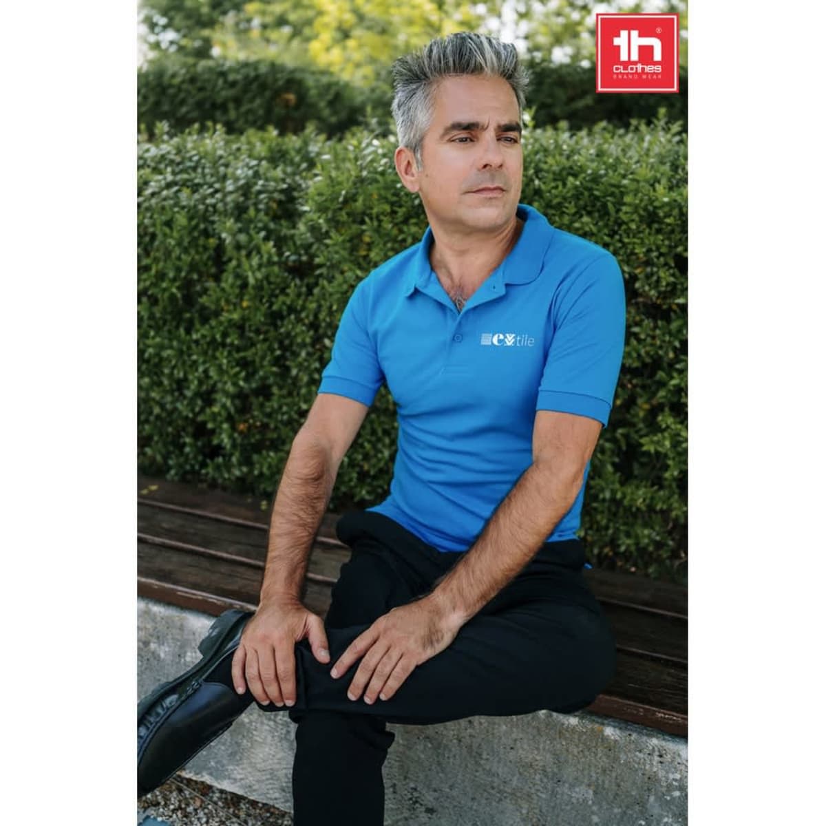 A blue men's polo shirt displayed on a man, styled with black trousers, seated outdoors. The image features a logo.