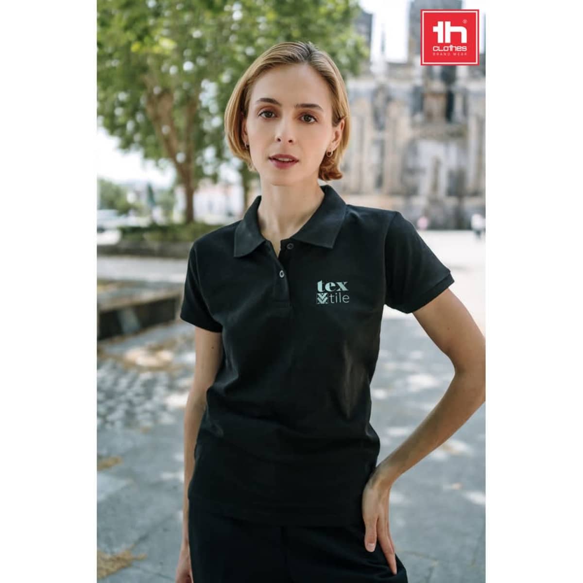 A woman wearing a black polo shirt with a logo, standing outdoors in an urban setting.