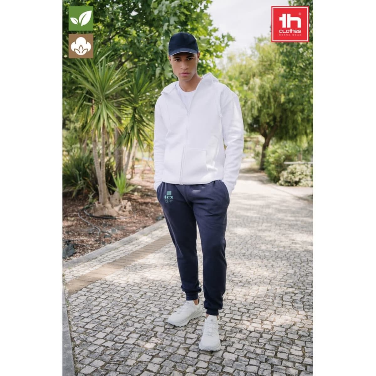 A model wearing navy unisex sweatpants with a white hoodie and a cap, standing in a landscaped area.
