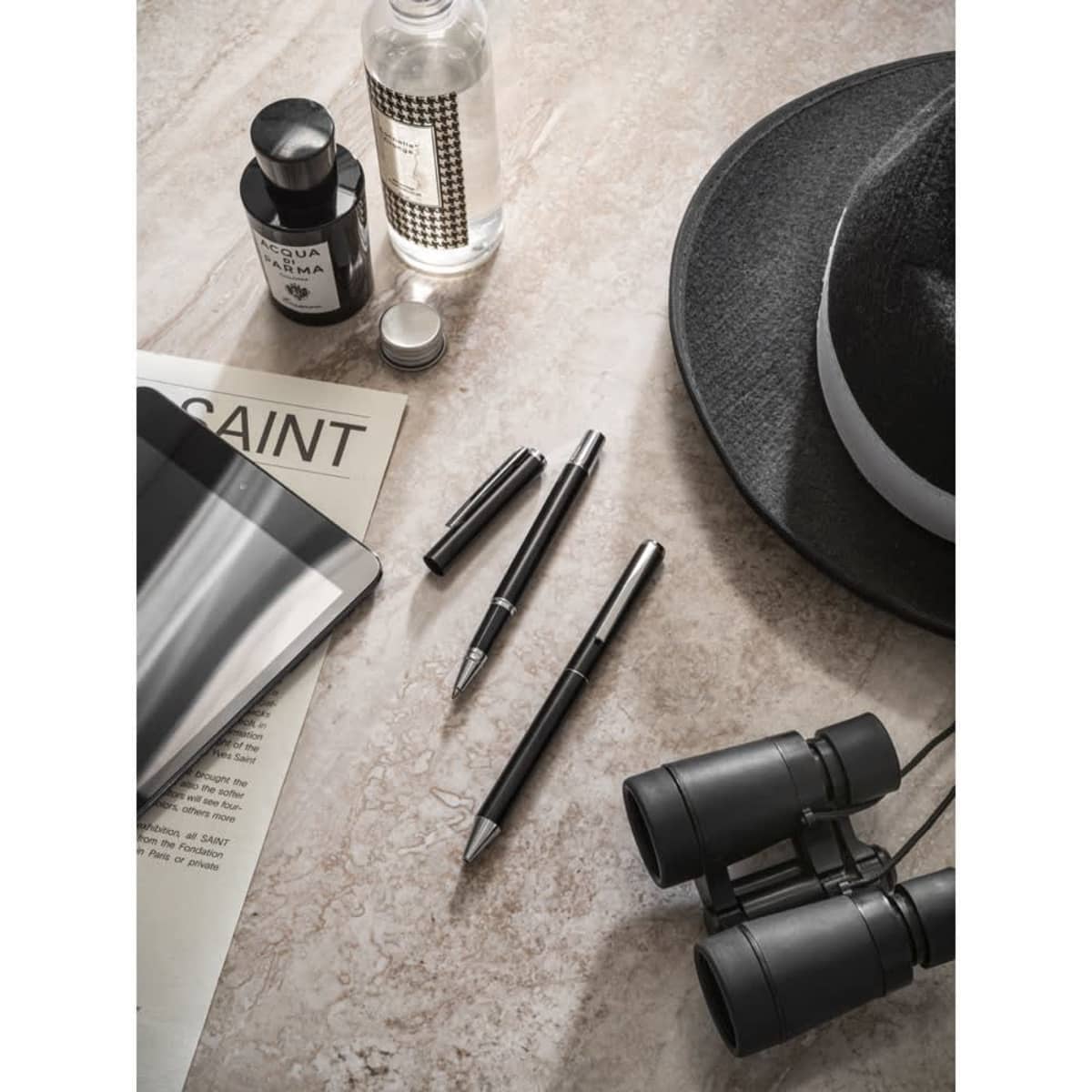 A roller and ballpoint pen set in aluminium, coloured silver and black, displayed with a notebook, hat, and binoculars.