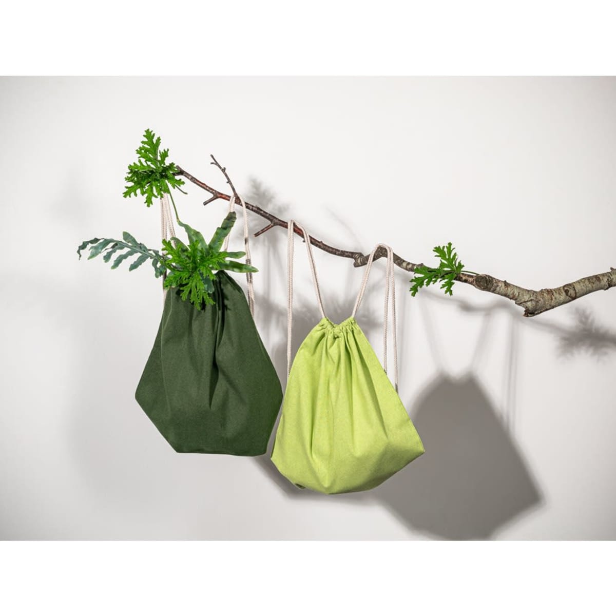 Two drawstring backpacks in dark green and light green materials hang from a branch, featuring a natural design backdrop.