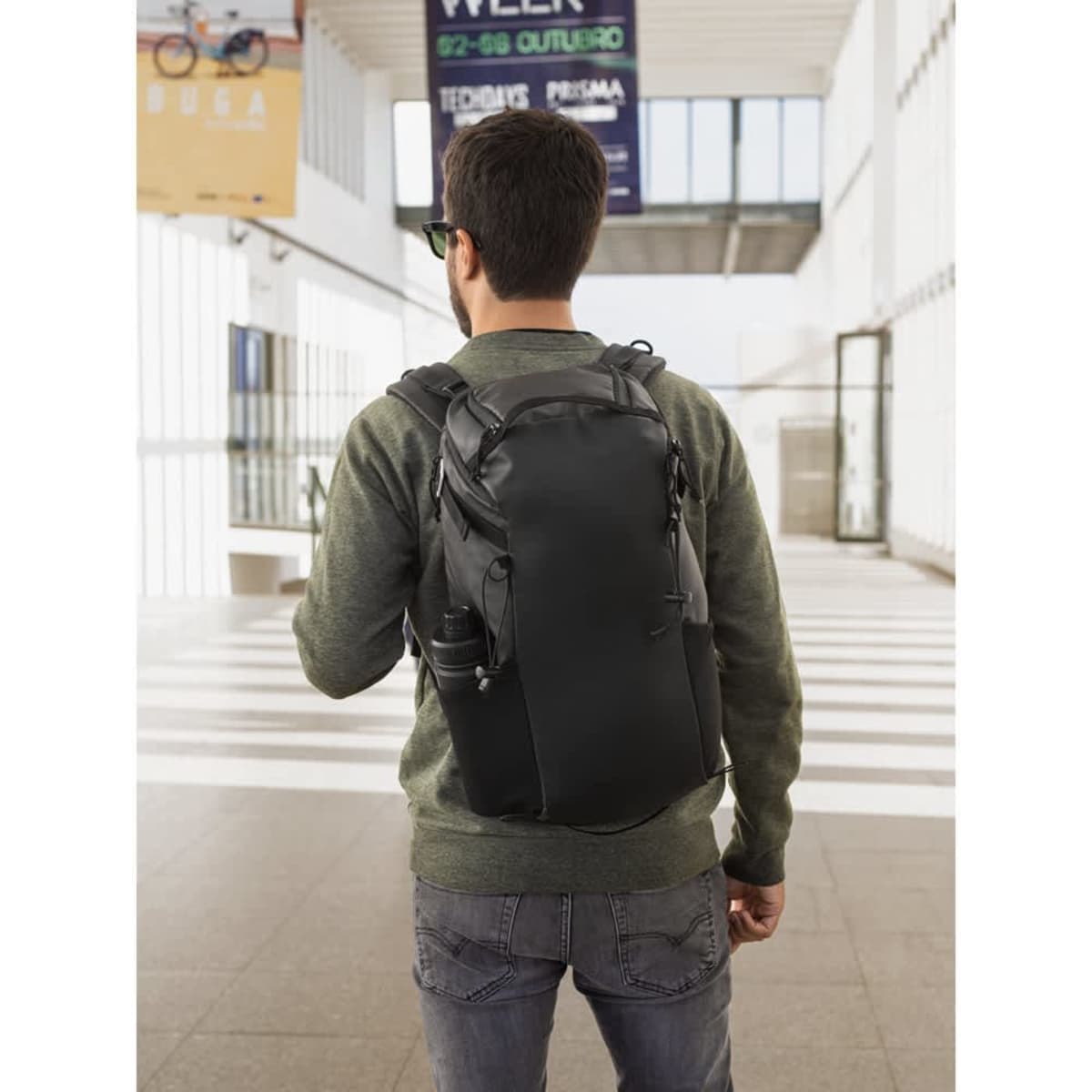 A black hiking backpack with a waterproof coating is worn by a person facing away, standing in a modern indoor space.