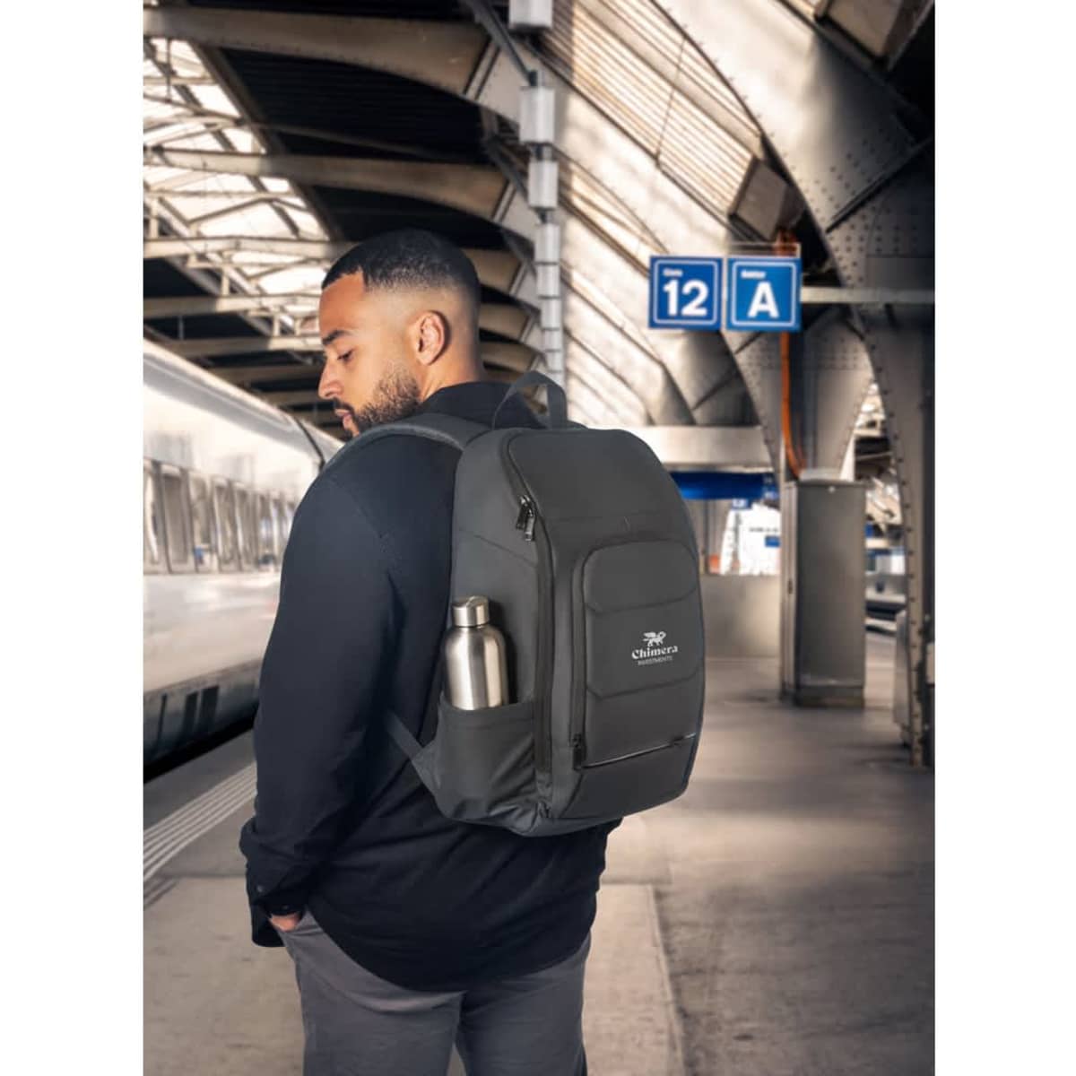 A black water-resistant notebook backpack with a drink bottle holder, displayed in a train station setting.