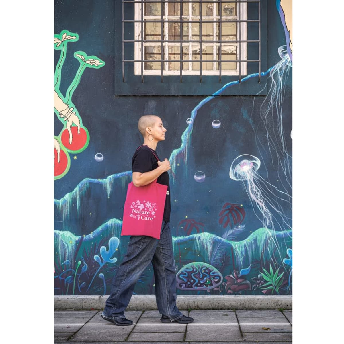 A person with a shaved head walks past a mural while carrying a pink tote bag made from recycled materials.