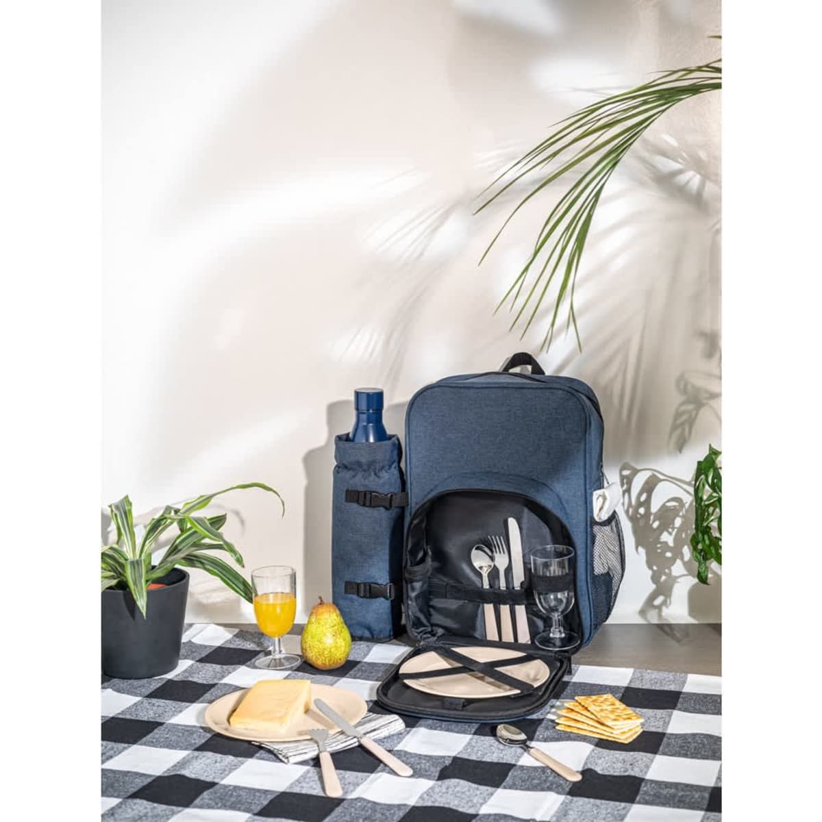 A navy picnic cooler backpack with cutlery, glasses, and a drink bottle, set on a checked tablecloth.