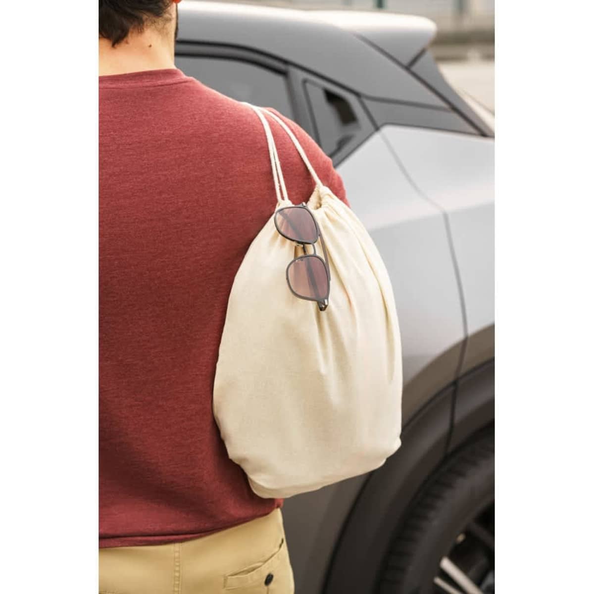 A cream drawstring bag with sunglasses hanging from it, being carried by a person near a vehicle.
