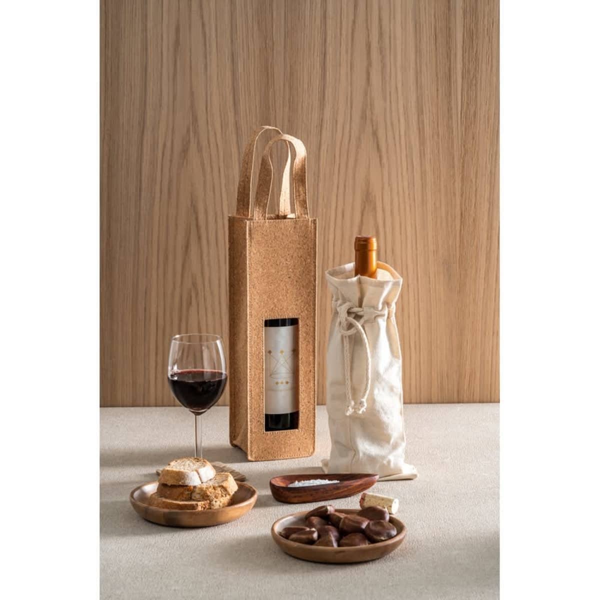 A drink bottle in a white cotton bag with a cork bag and a glass of red wine on a table alongside chocolates and biscuits.