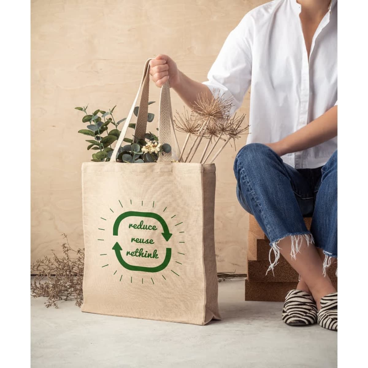 A natural-toned tote bag with two handles, featuring a logo and greenery inside, displayed by a person in casual attire.