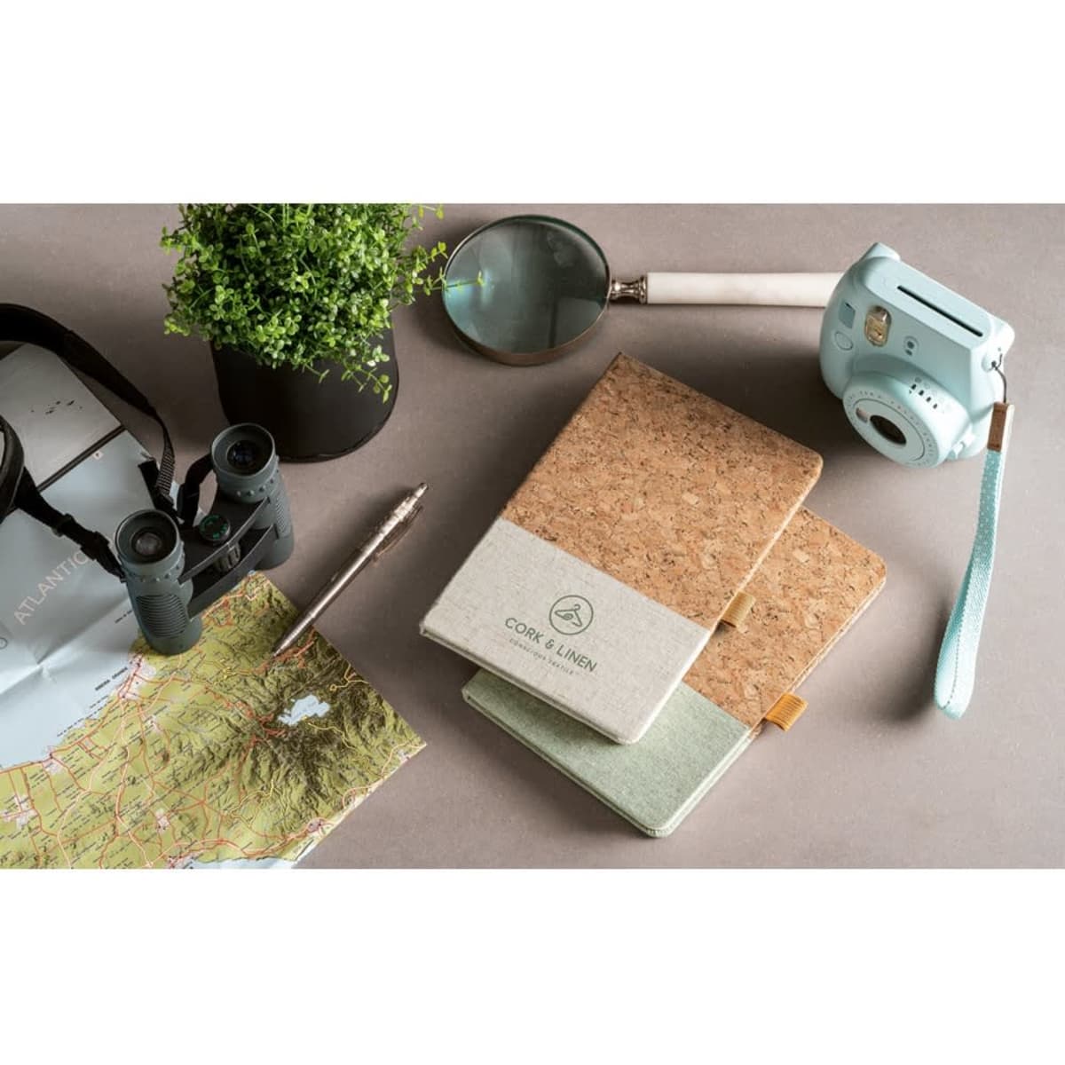 A cork and linen A5 notebook in green and beige, placed alongside binoculars, a magnifying glass, and a camera.