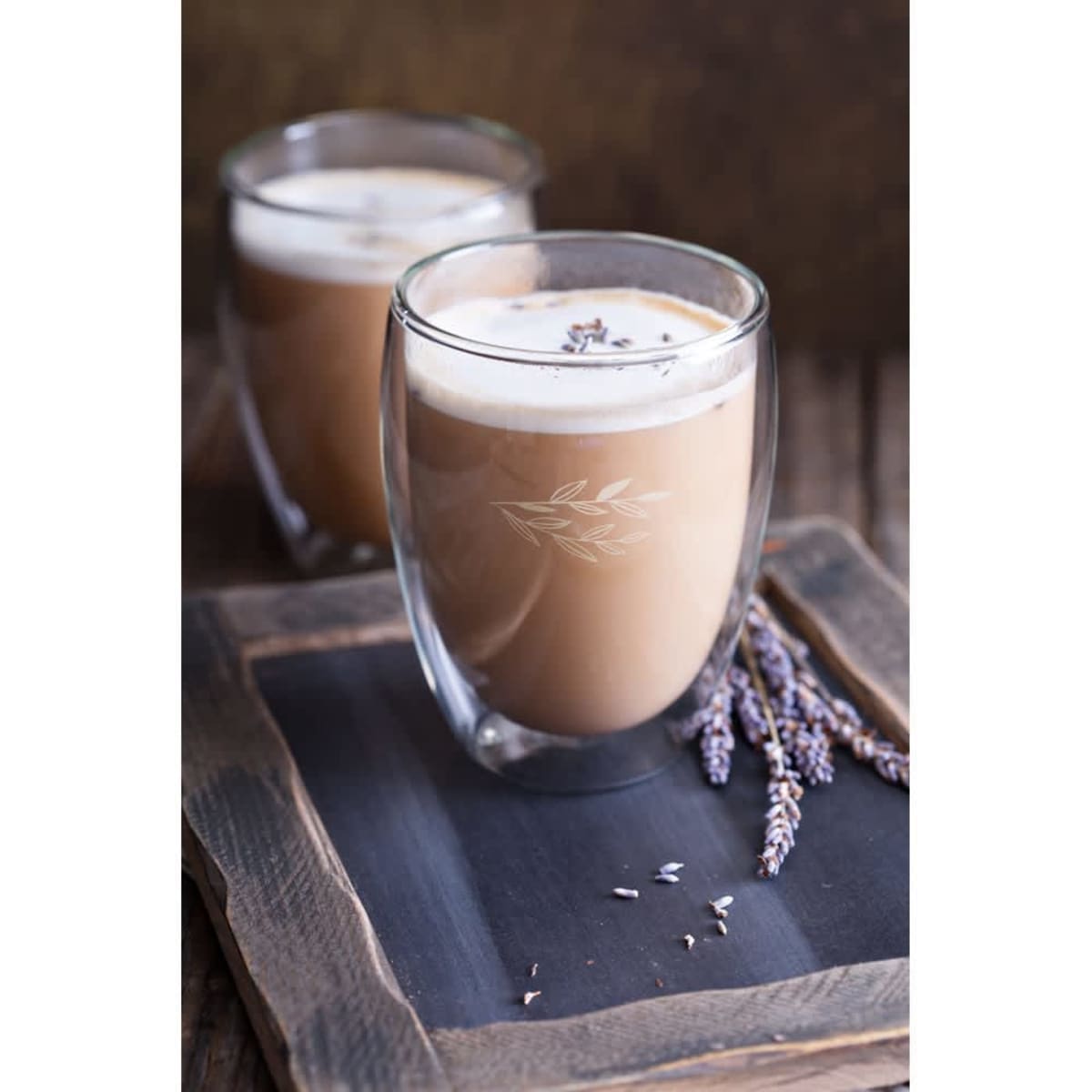 A pair of clear borosilicate glass cups filled with coffee, featuring frothy cream and garnished with lavender.