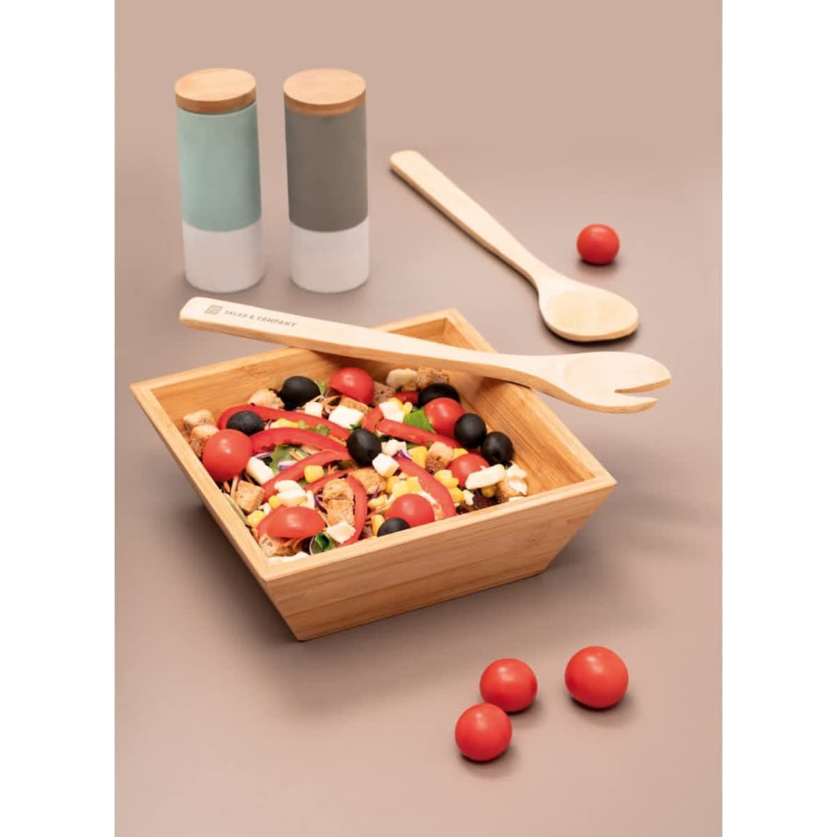 A square bamboo salad bowl filled with colourful ingredients, accompanied by wooden utensils and two salt shakers.