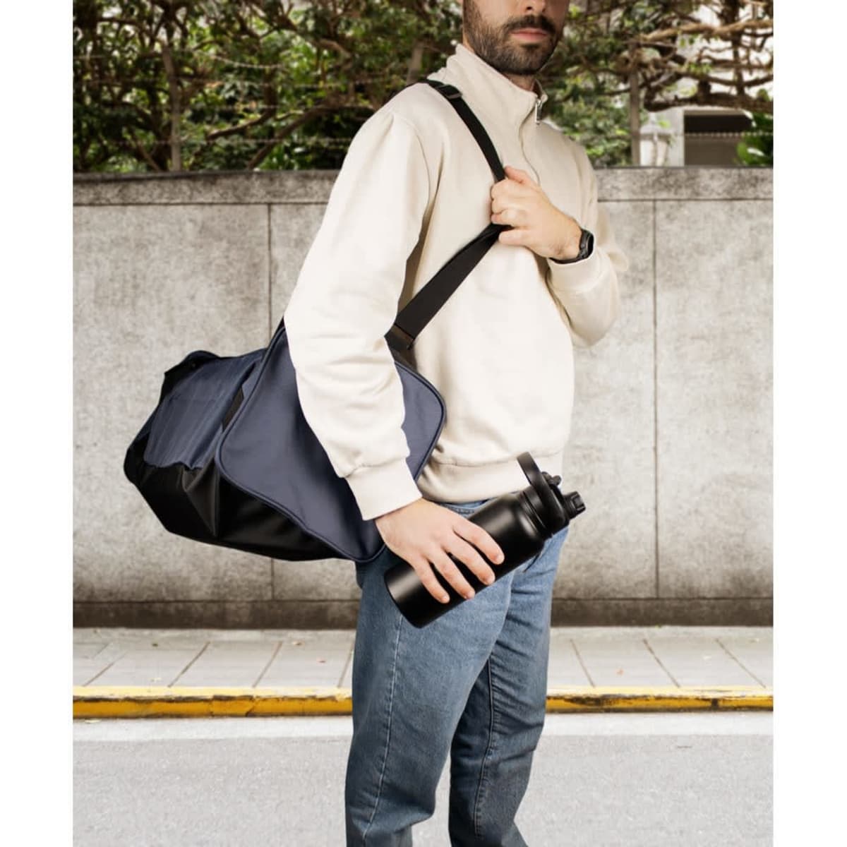A drink bottle in matte black, made of stainless steel, with a double wall vacuum insulation, held by a person beside a bag.