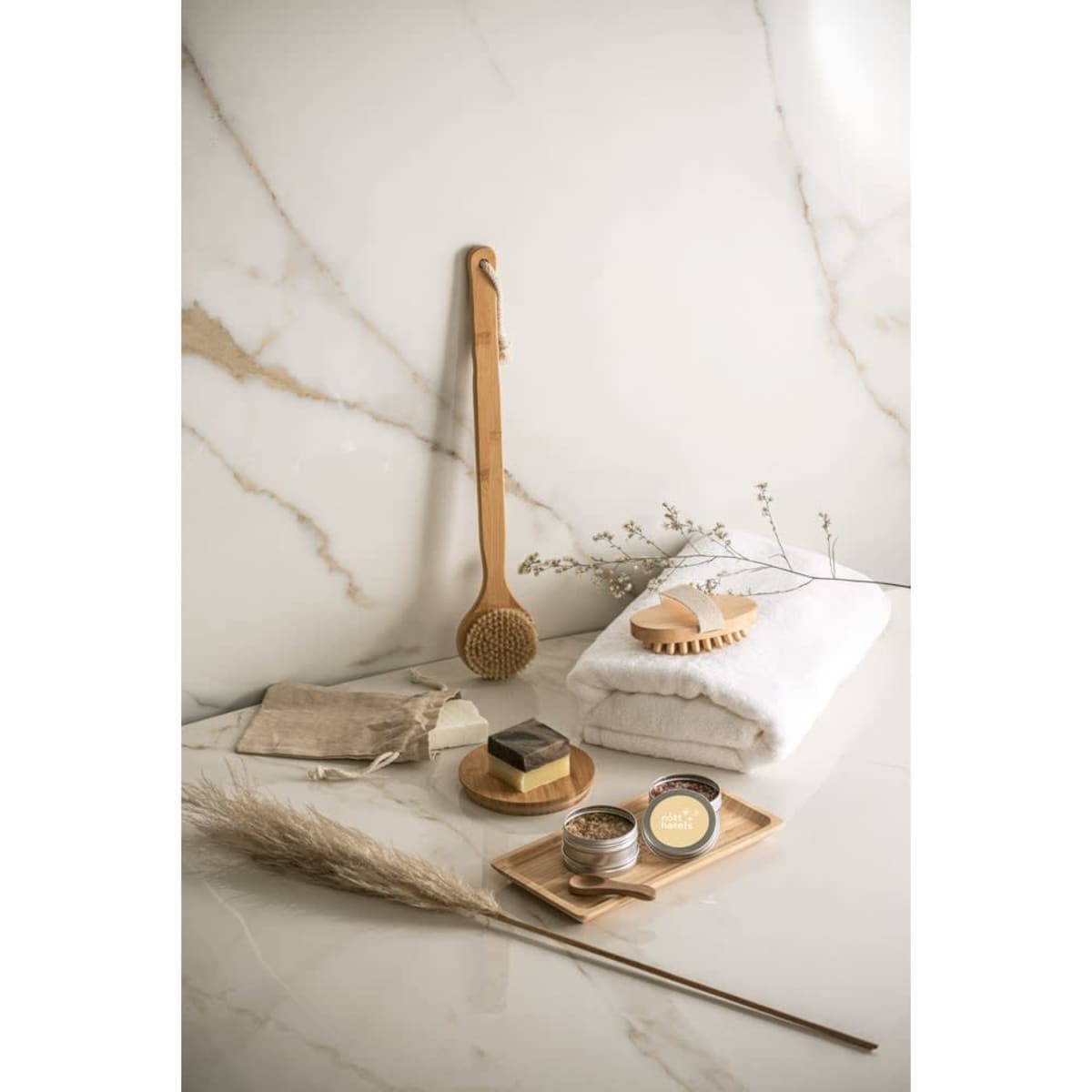 A bamboo shower and bath brush rests on a marble surface, surrounded by towels, jars, and natural decor.