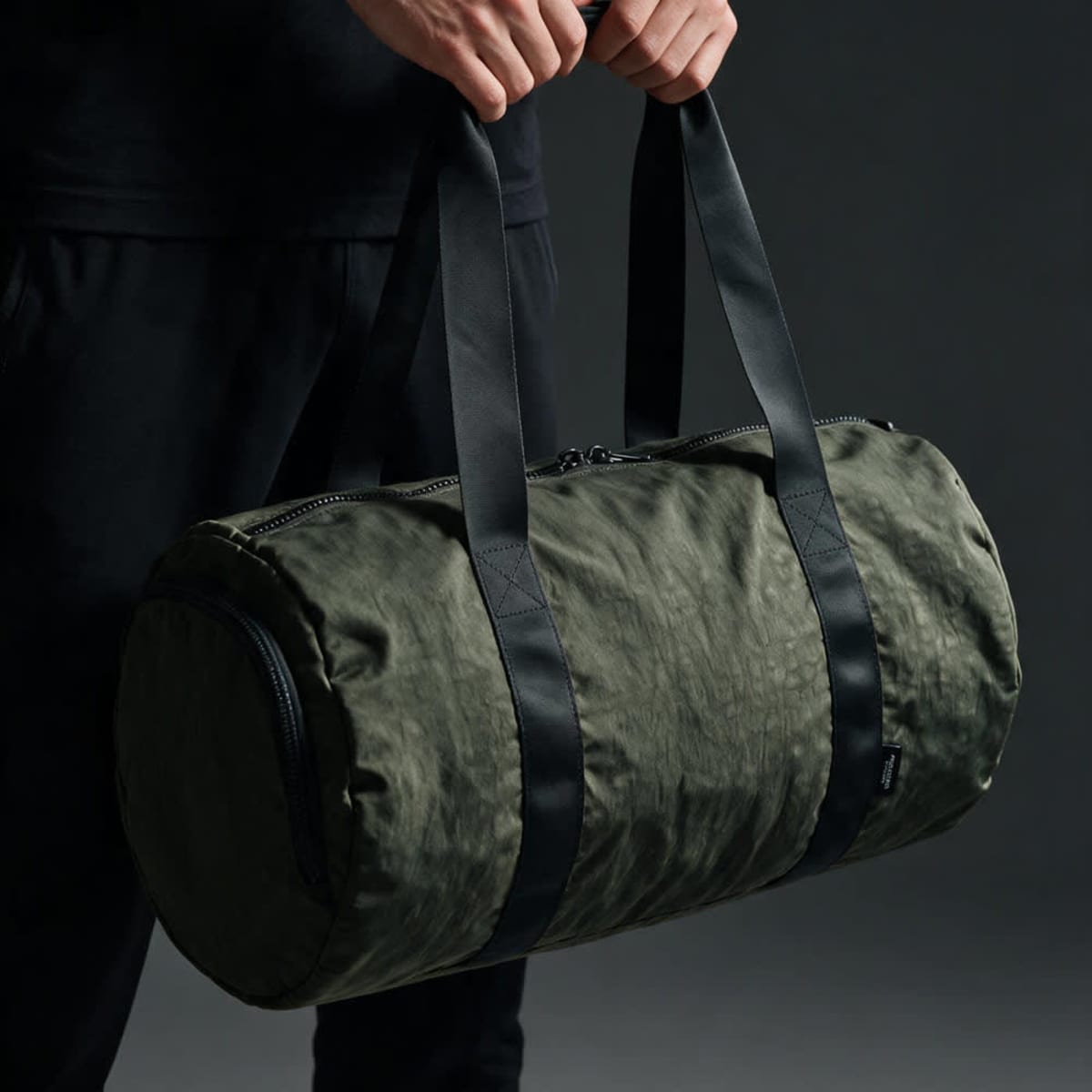 A green gym bag with black straps and a zip closure, held by a person. It features a rounded design and a branding logo.