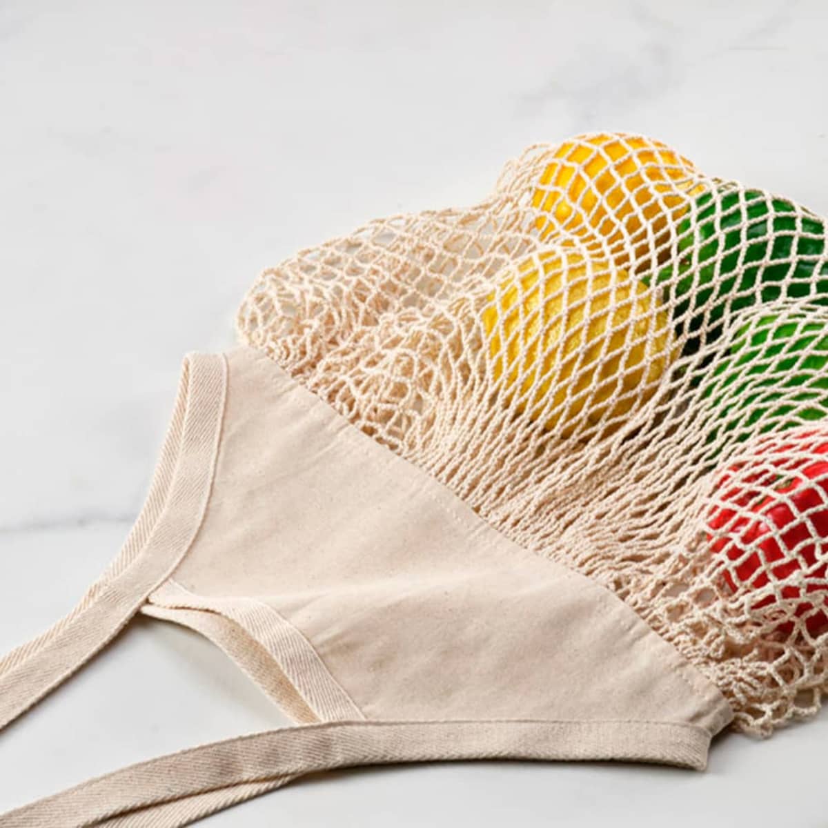 A beige vegetable bag made of cotton, featuring a netted design with various coloured fruits visible inside.