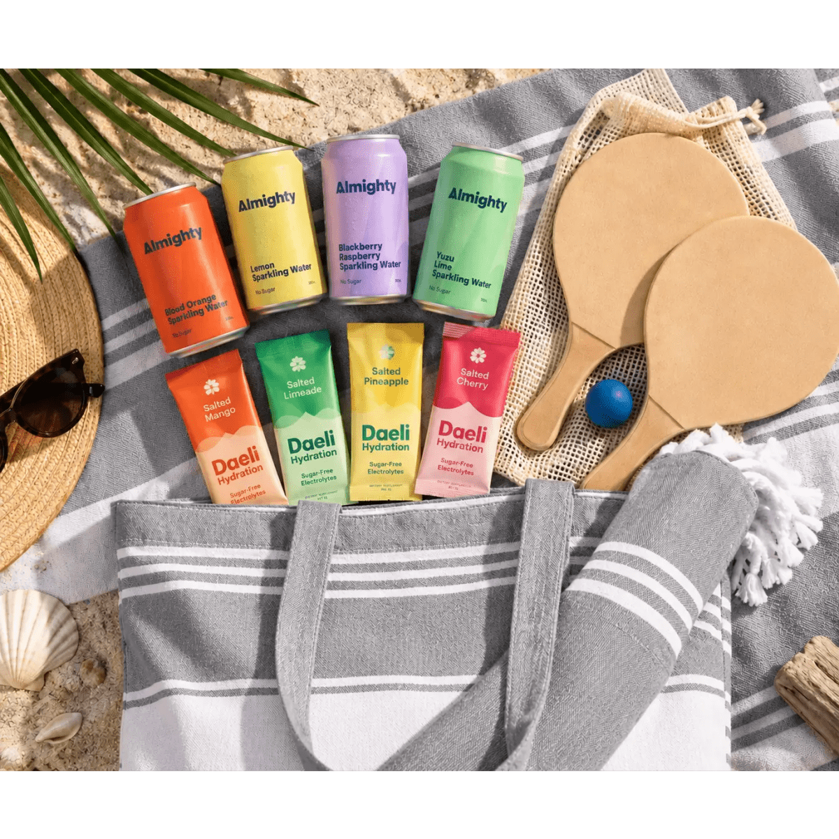 A grey and white tote bag holds various drinks and two wooden paddle bats, arranged on a beach-themed blanket.