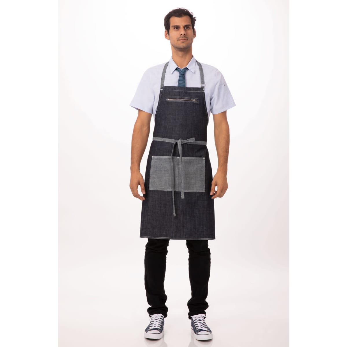 A man wearing a dark denim bib apron with grey accents and pockets, paired with a light shirt and tie.
