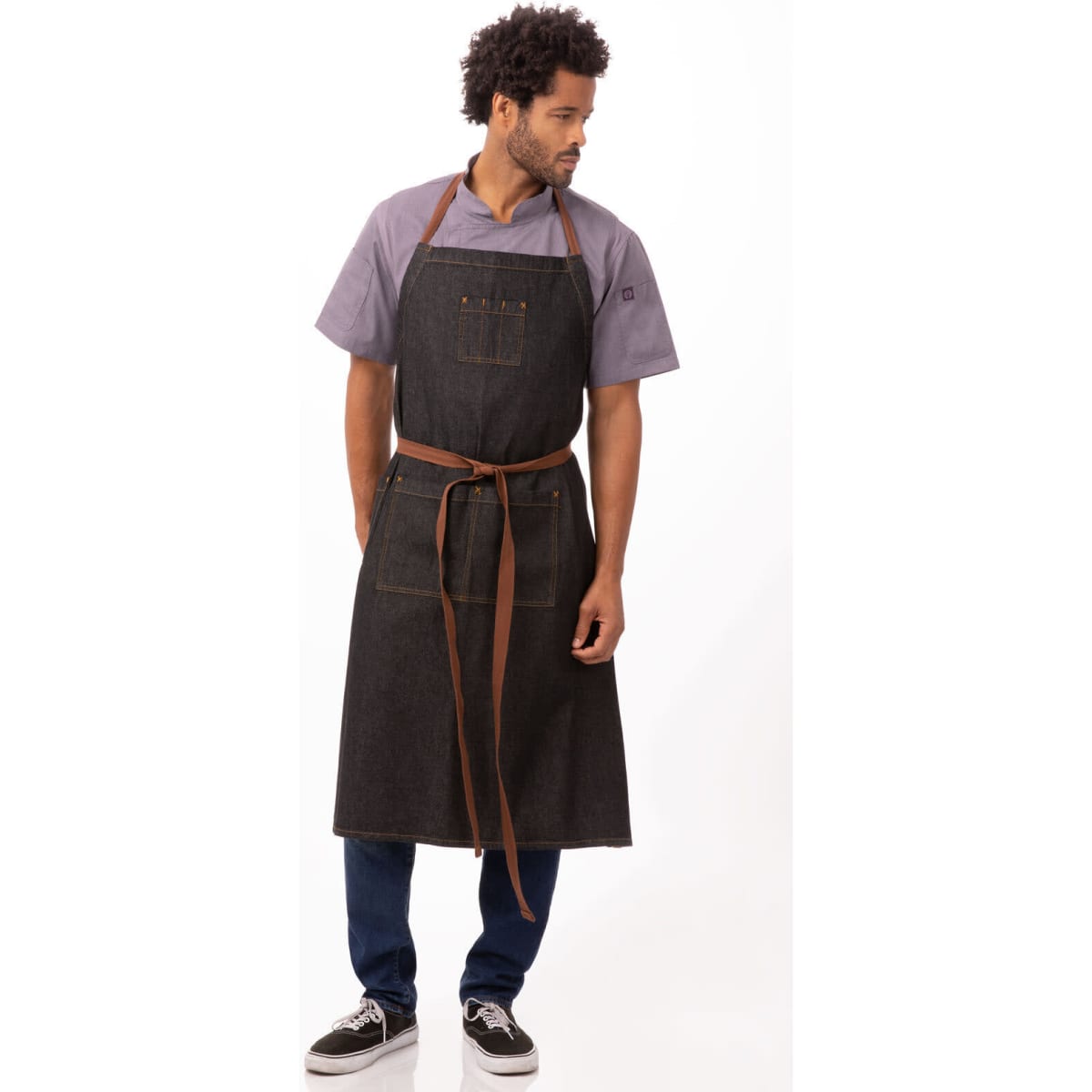 A dark denim apron with brown straps, featuring pockets and worn by a model standing gracefully.