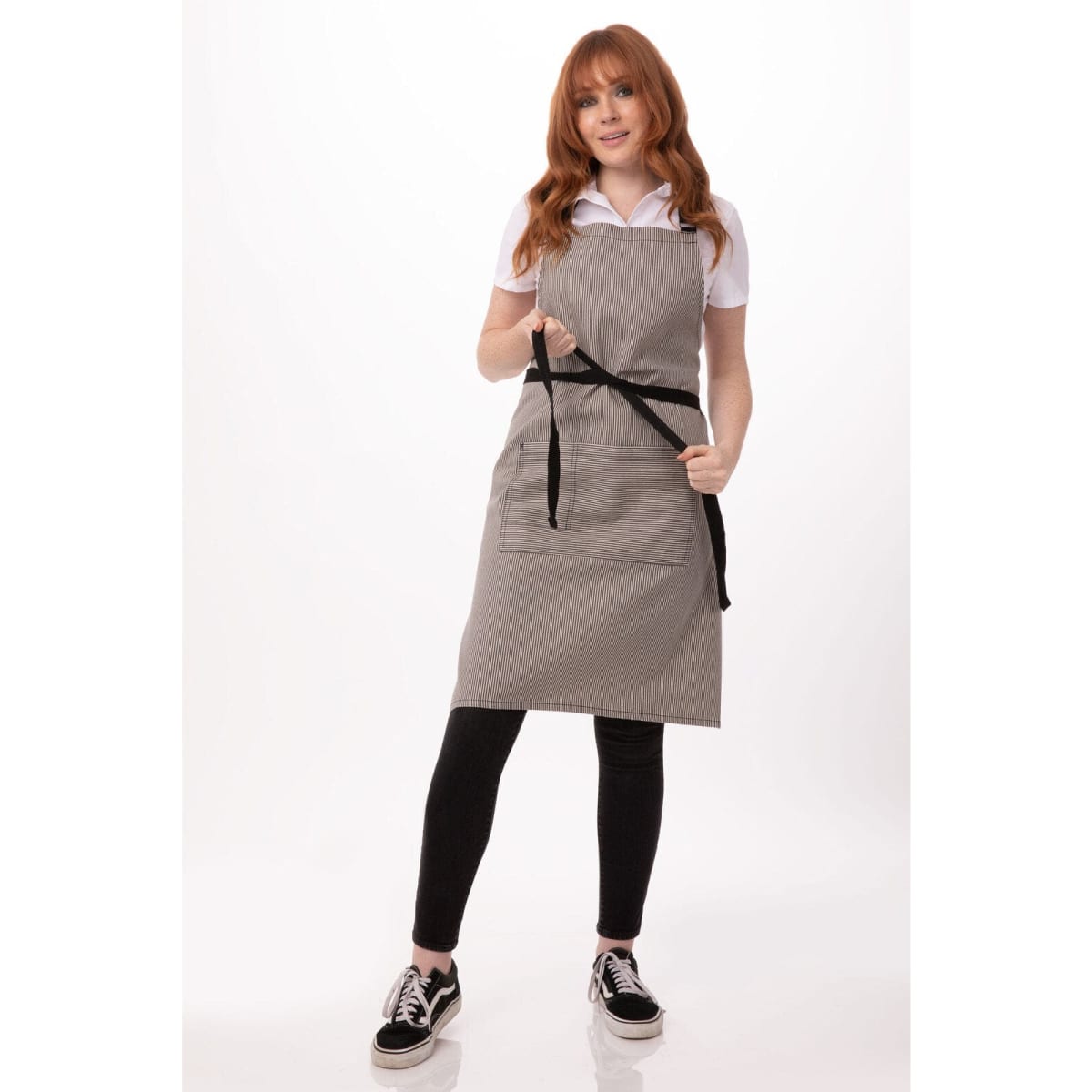 A woman wearing a grey Portland Bib Apron with black straps, standing confidently and holding the apron ties.