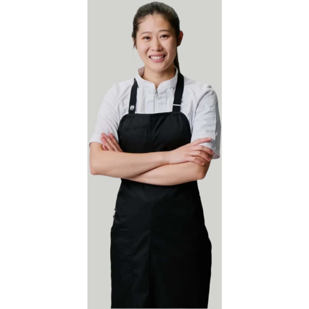 A person wearing a black bib apron over a white shirt, arms crossed, smiling against a neutral background.