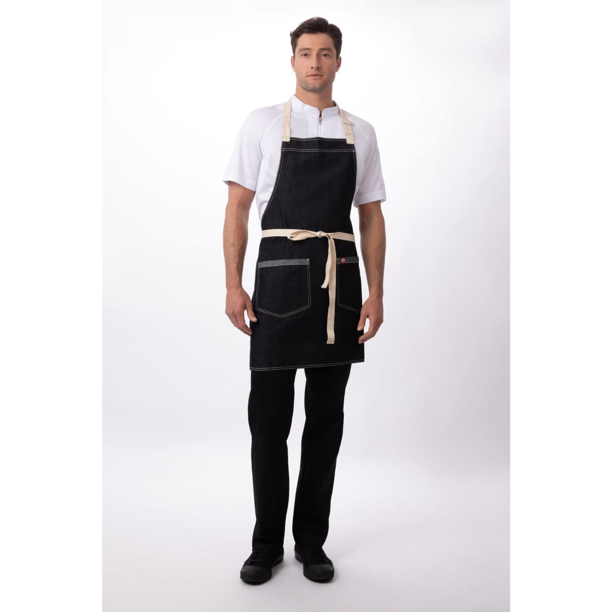 A man wearing a black Ridgewood Bib Apron with a beige tie and pockets, standing against a white background.