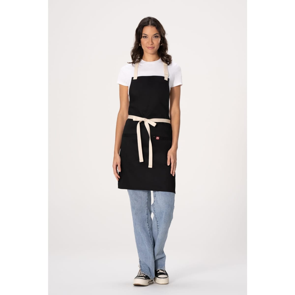 A woman wearing a black Ridgewood Bib Apron with a cream-coloured tie, paired with casual jeans and a white t-shirt.