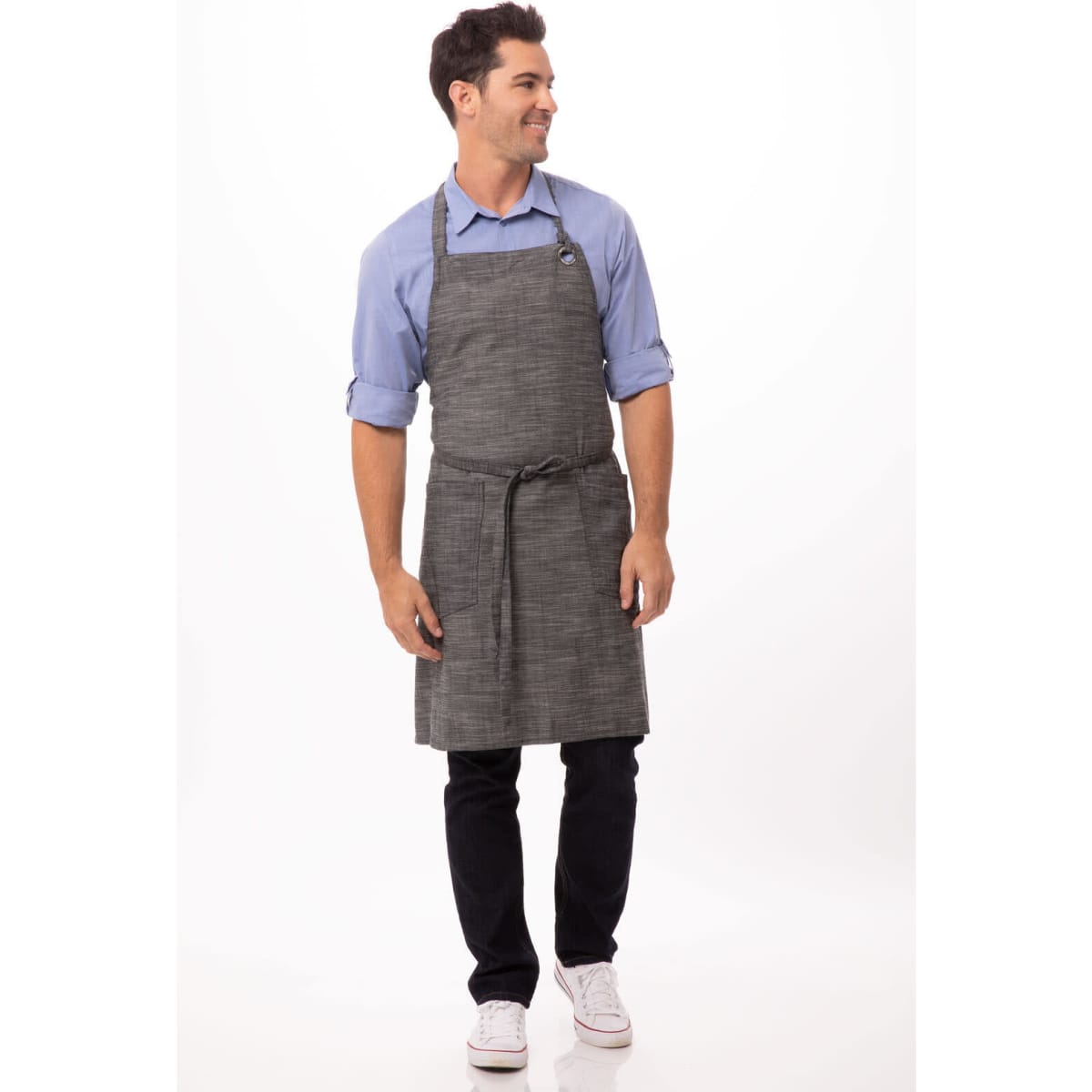 A man wearing a grey bib apron over a blue shirt, styled with black trousers and white shoes. The apron features a logo.