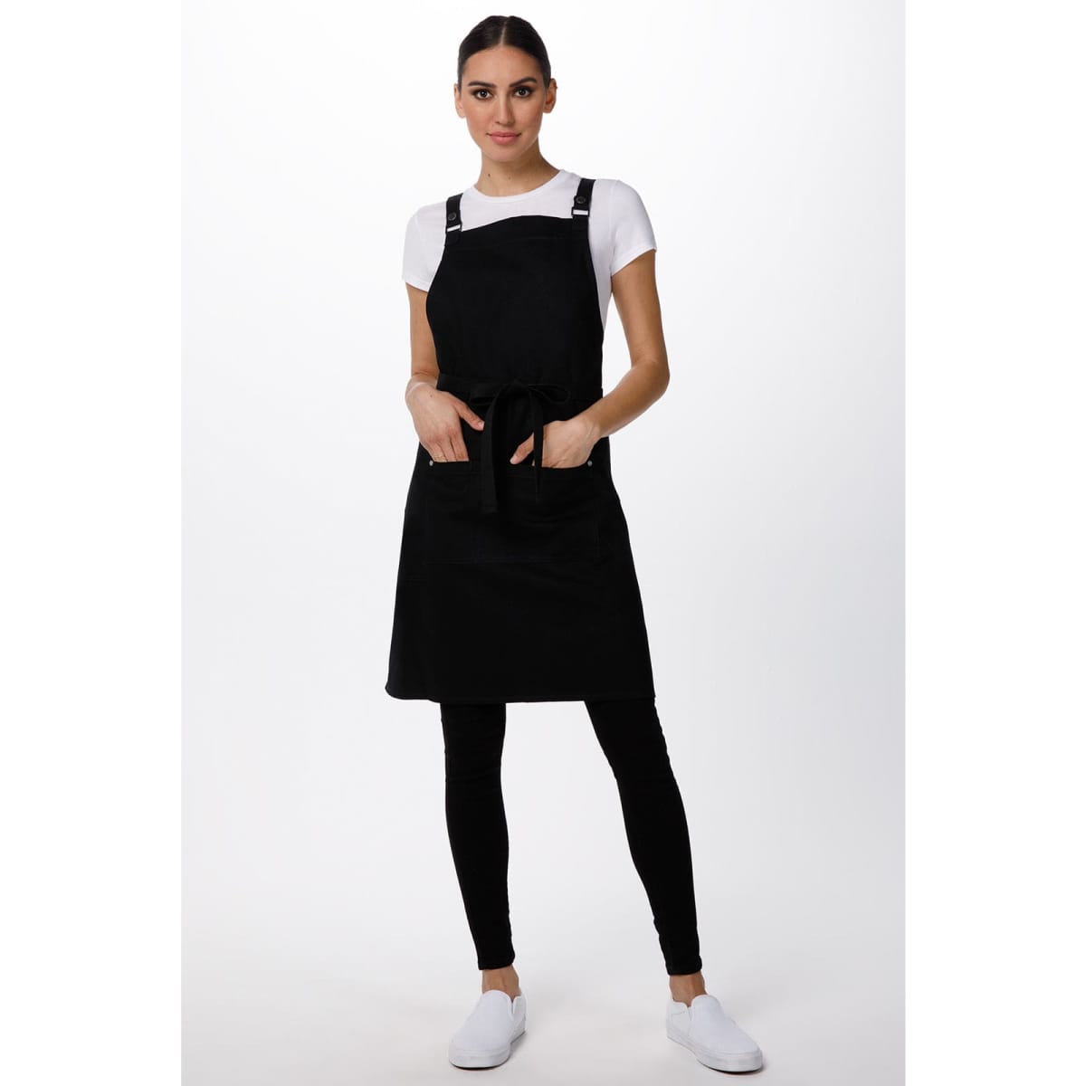 A black cross-back apron with pockets, worn over a white T-shirt and black leggings, featuring branding.