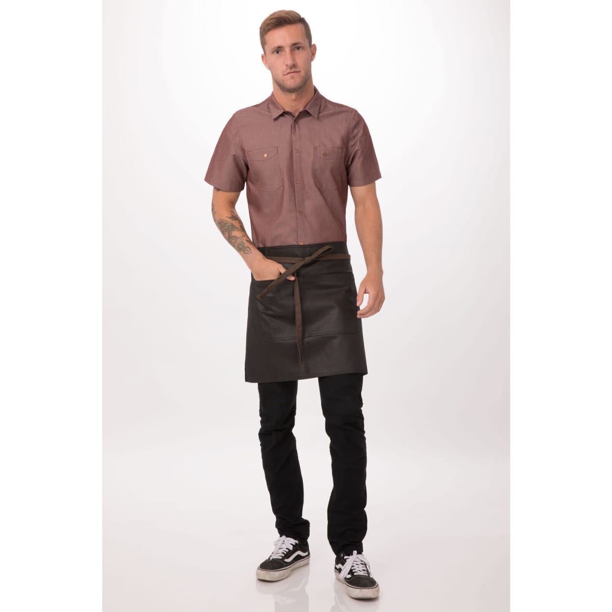 A man wearing a brown shirt and a black half apron with a waist tie, standing against a light background.