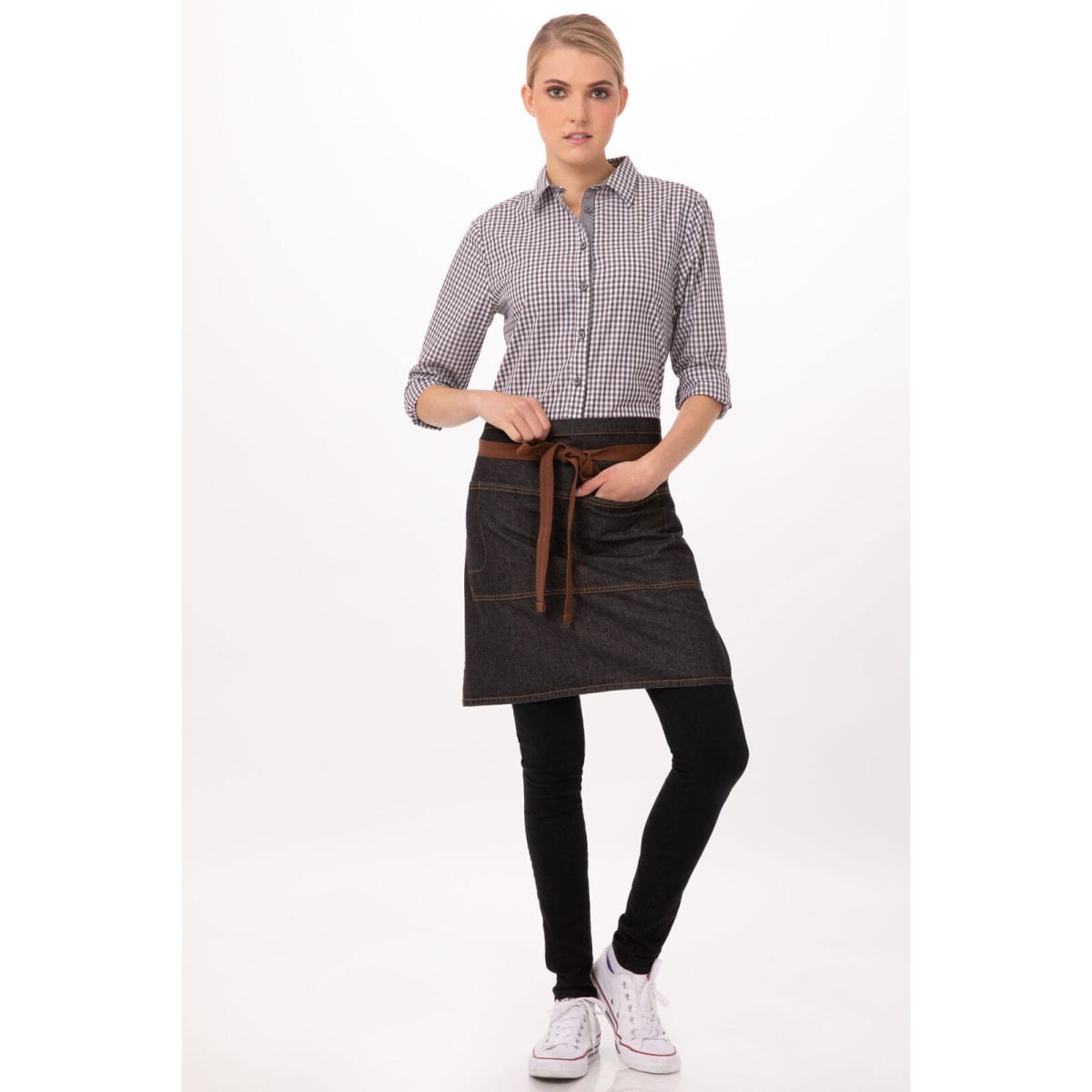 A woman wearing a stylish black and denim half bistro apron with a brown tie, paired with a checkered shirt and black pants.