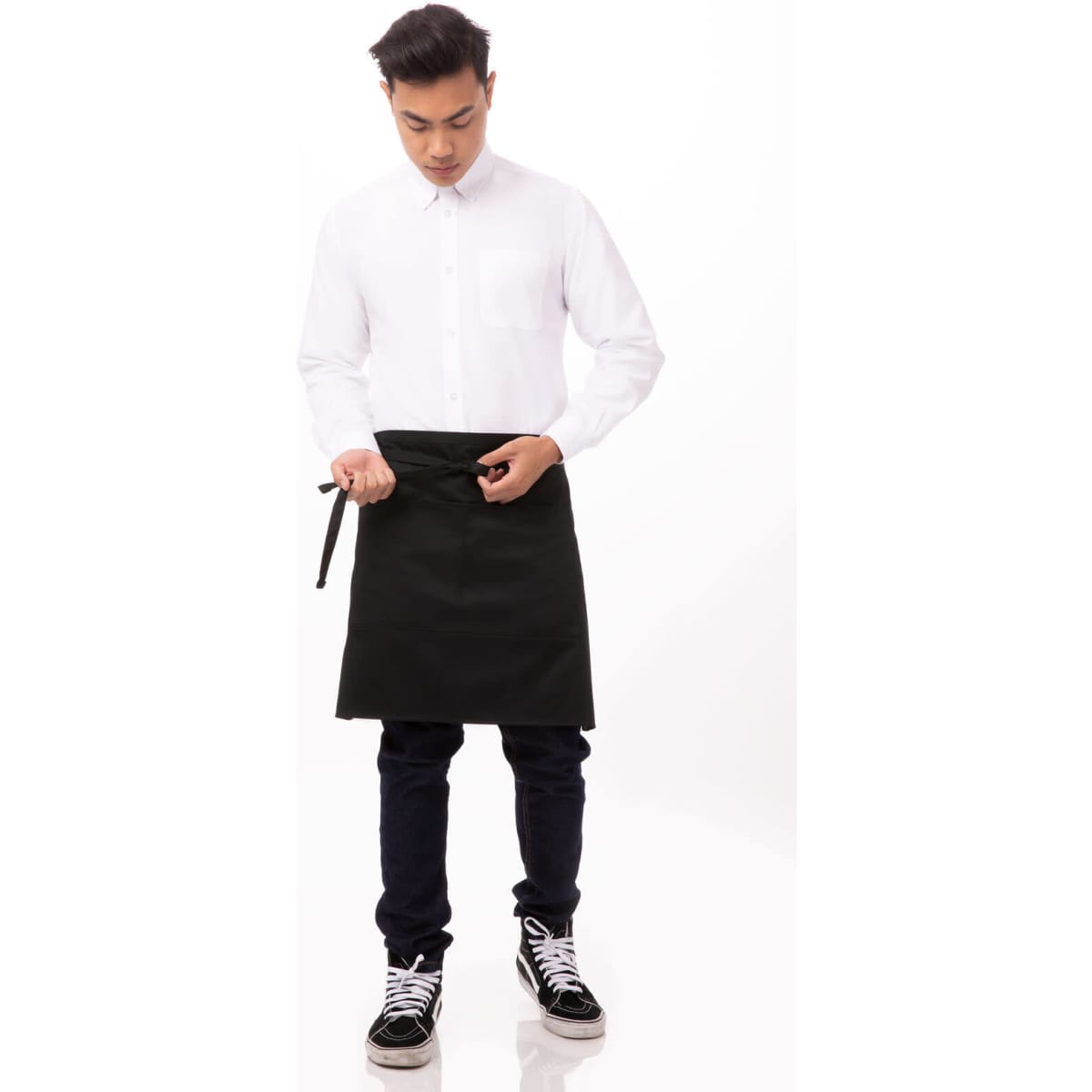 A half bistro apron in black, worn by a person adjusting the ties, paired with a white shirt and dark jeans.