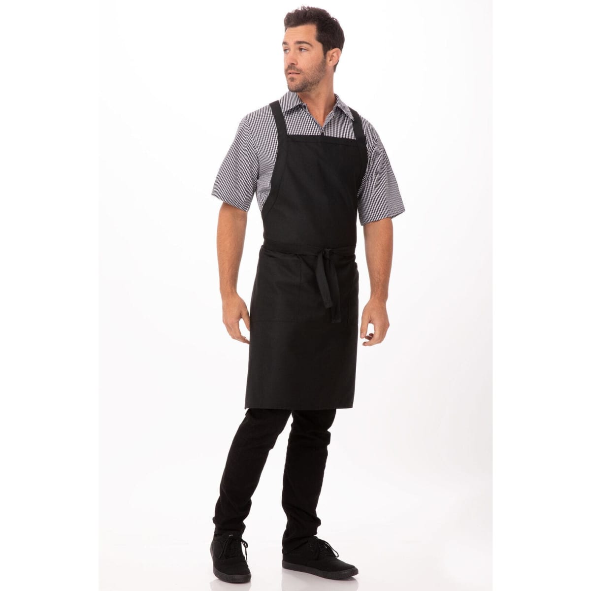 A man wears a black cross-back bib apron over a patterned shirt and black pants, with one hand on his hip.