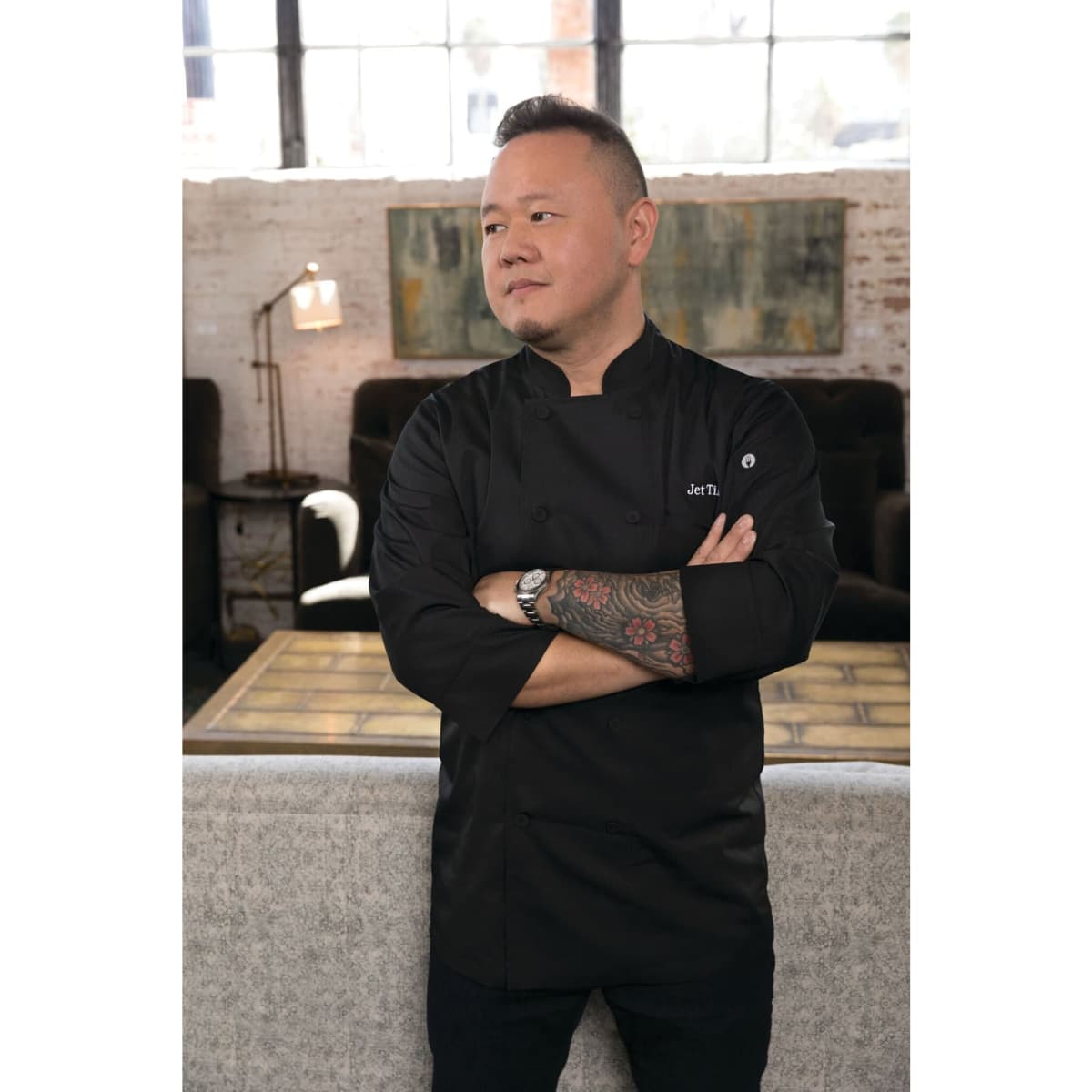 A chef wearing a black jacket stands with arms crossed, showcasing a tattooed arm and a stylish interior background.