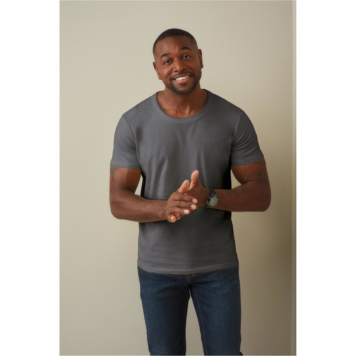 A man wearing a grey crew neck t-shirt and blue jeans, smiling and gesturing with his hands.