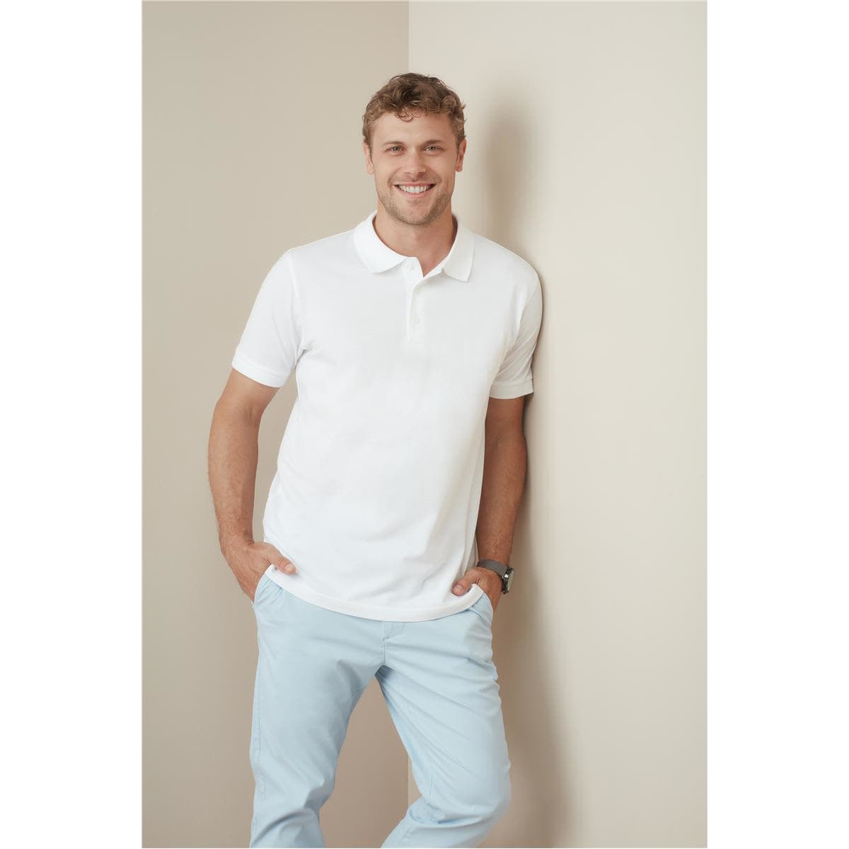 A man wearing a white premium cotton polo shirt, smiling while leaning against a wall, paired with light blue trousers.